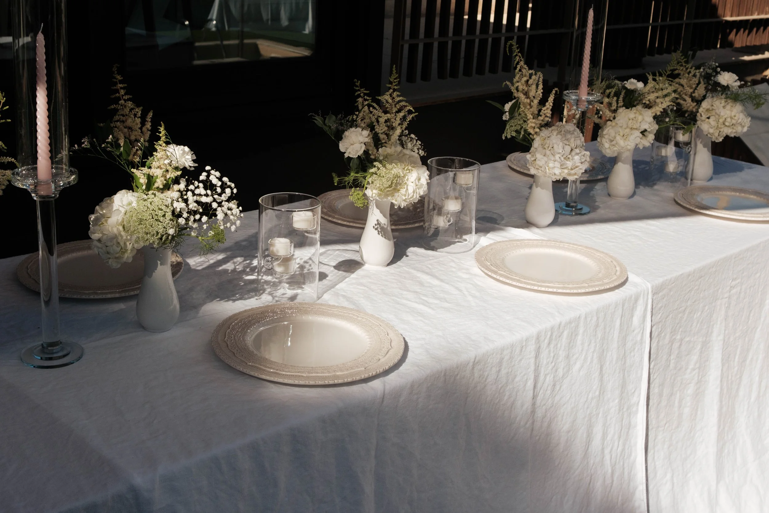 Ein festlich gedeckter Tisch mit weißen Vasen mit weißen Blumen, Kerzen in Gläsern, weißen Tellern, auf der rechten Seite sind zahlreiche Blumenarrangements, die den Tisch schmücken.
