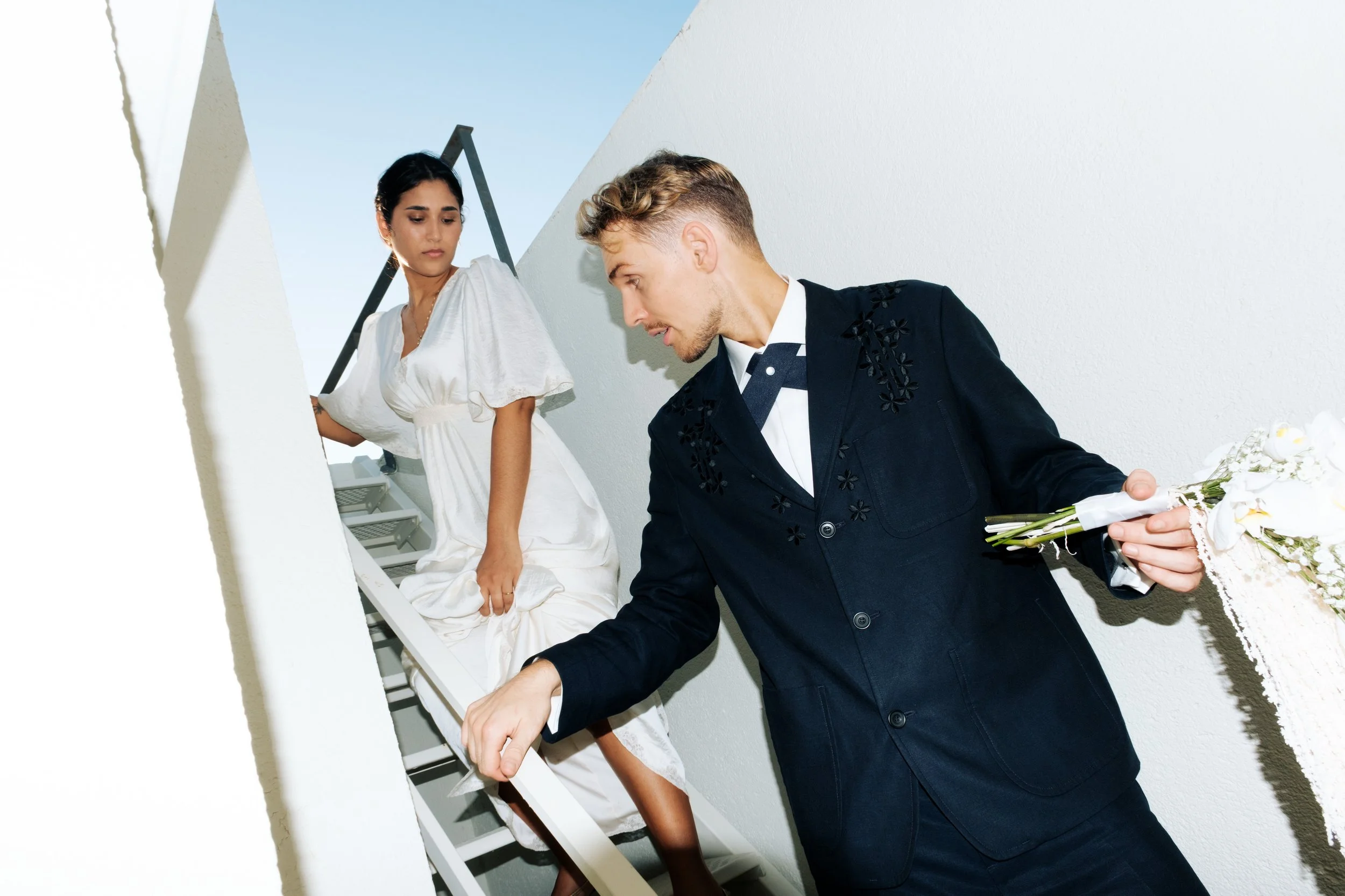 Ein Mann in einem schwarzen Anzug mit Blumenmuster hält einen Blumenstrauß, während eine Frau in einem weißen Kleid die Treppe hochsteigt.