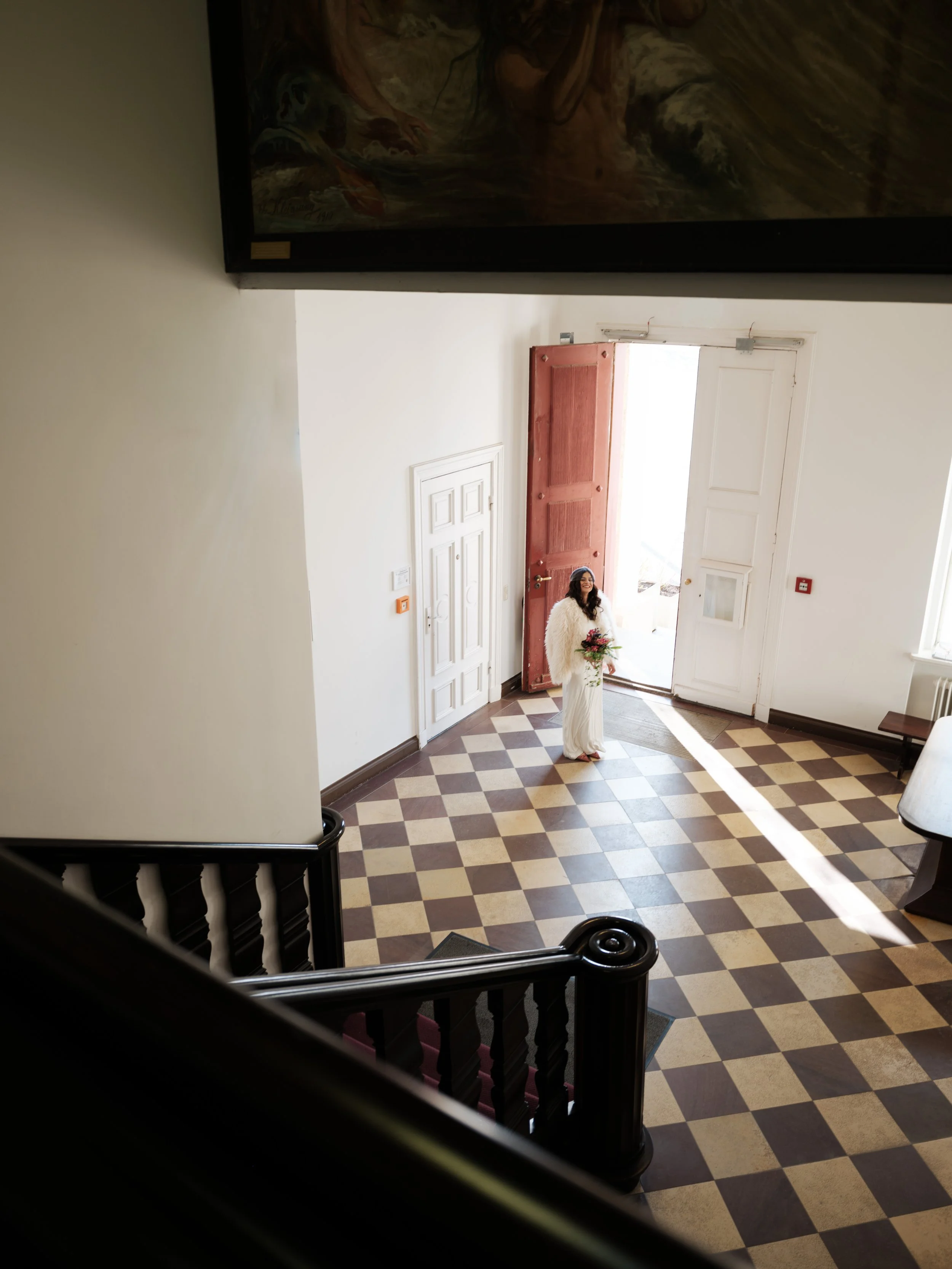 Eine junge Frau in einem weißen Kleid mit einem Blumenstrauß steht in einem hellen, eleganten Eingangsbereich vor einem offenen roten Tür. Sie lächelt und ist von Sonnenlicht umgeben.