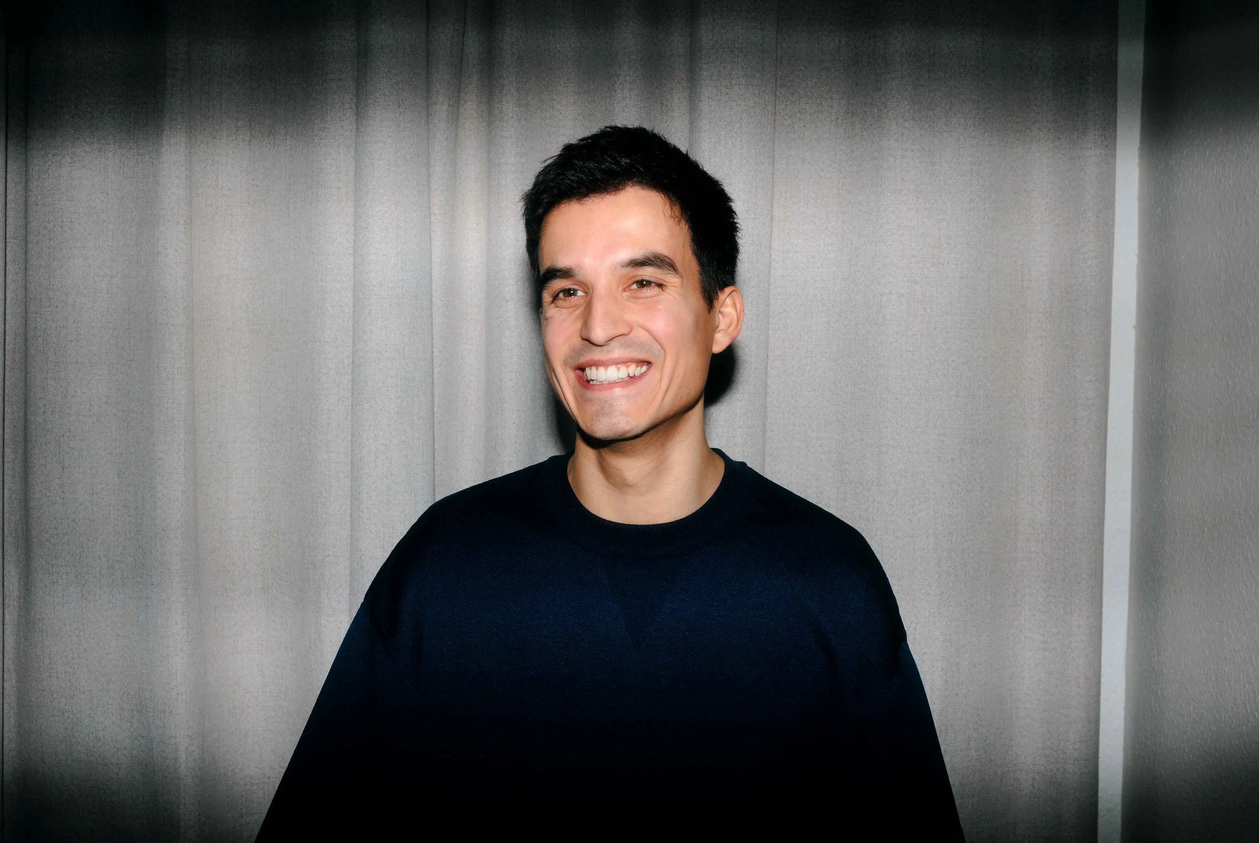 Lächelnder Mann mit dunklem Haar vor grauer Kulisse.