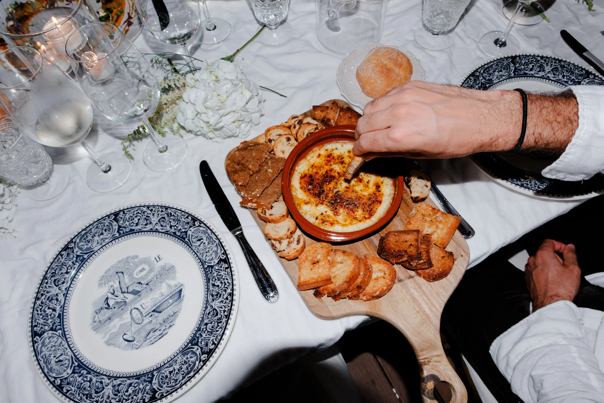 Ein gedeckter Tisch mit weißen Tischtüchern, mehreren leeren Weingläsern, einer Blumenarrangement und einem Baguette. In der Mitte steht ein Holzbrett mit einer Keramikschüssel, aus der Käse oder Aufstrich mit geröstetem Brot serviert wird. Eine Hand