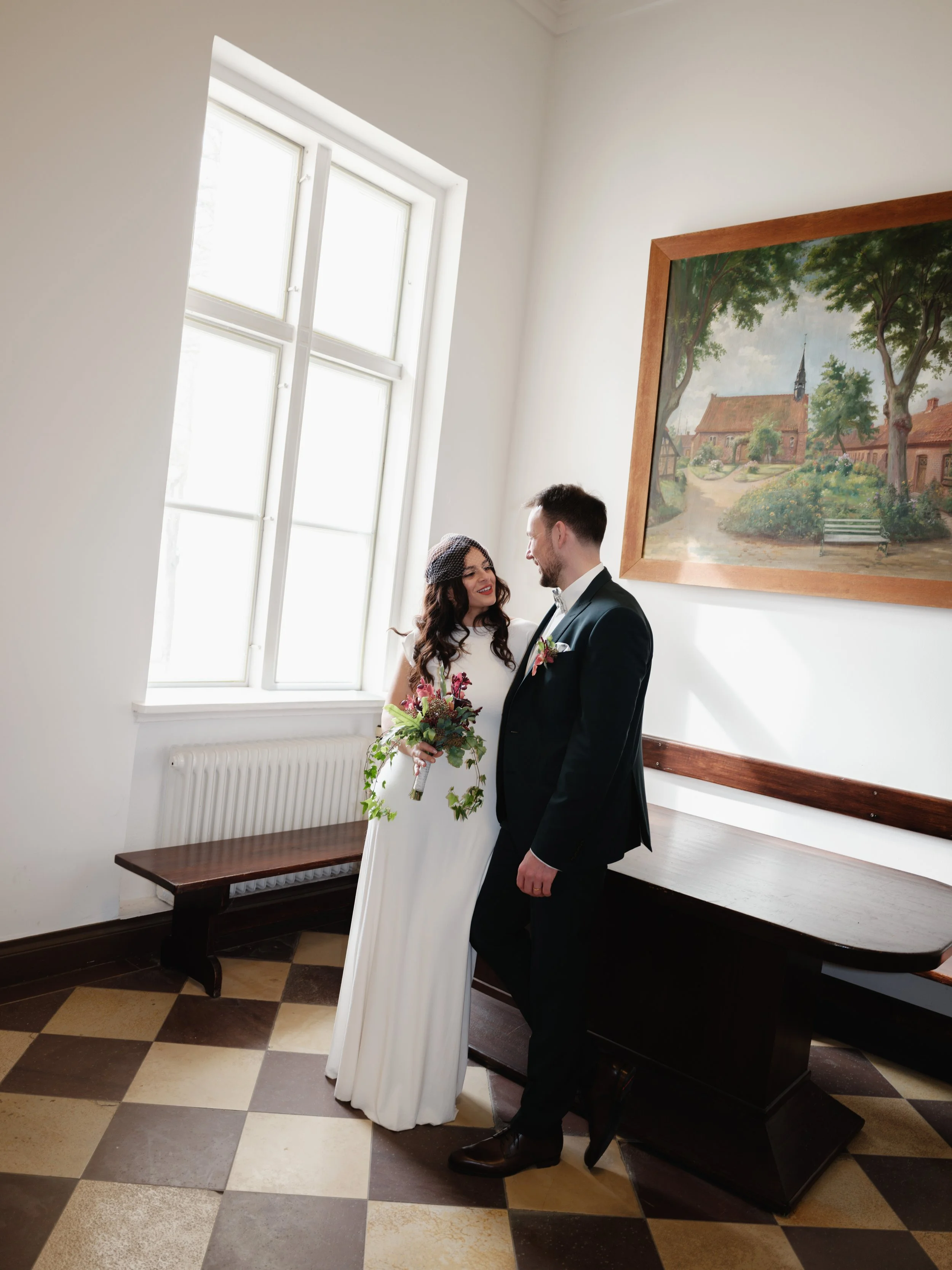Ein Brautpaar steht in einem hellen Raum nebeneinander, die Braut hält einen Blumenstrauß, beide lächeln sich an. Im Hintergrund sind ein Fenster, ein Bild und eine Bank sichtbar.