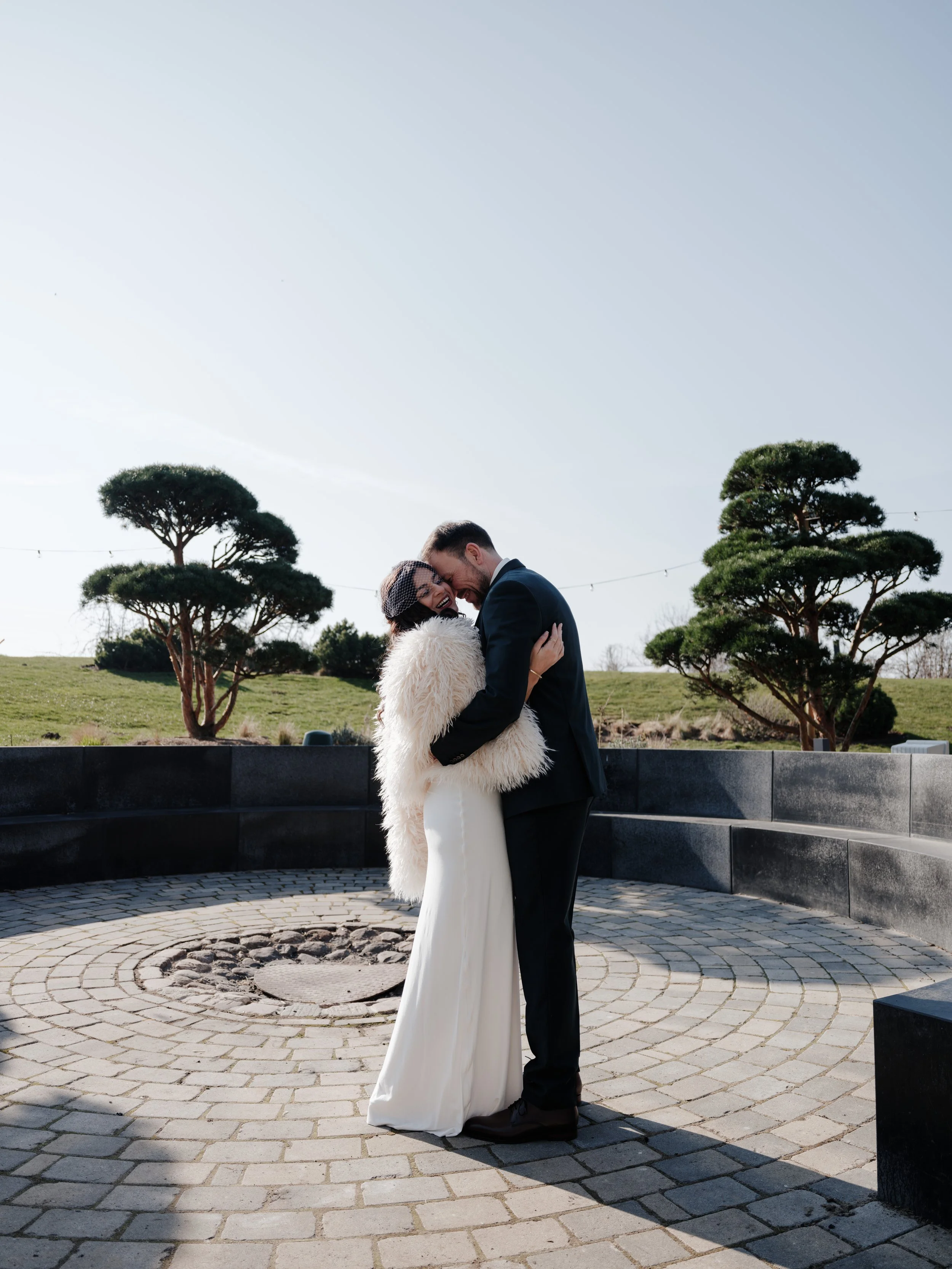 Ein Paar in formeller Kleidung umarmt sich auf einer Steinbodenfläche im Freien, umgeben von grünem Rasen und geometrisch geschnittenen Bäumen, bei sonnigem Himmel.