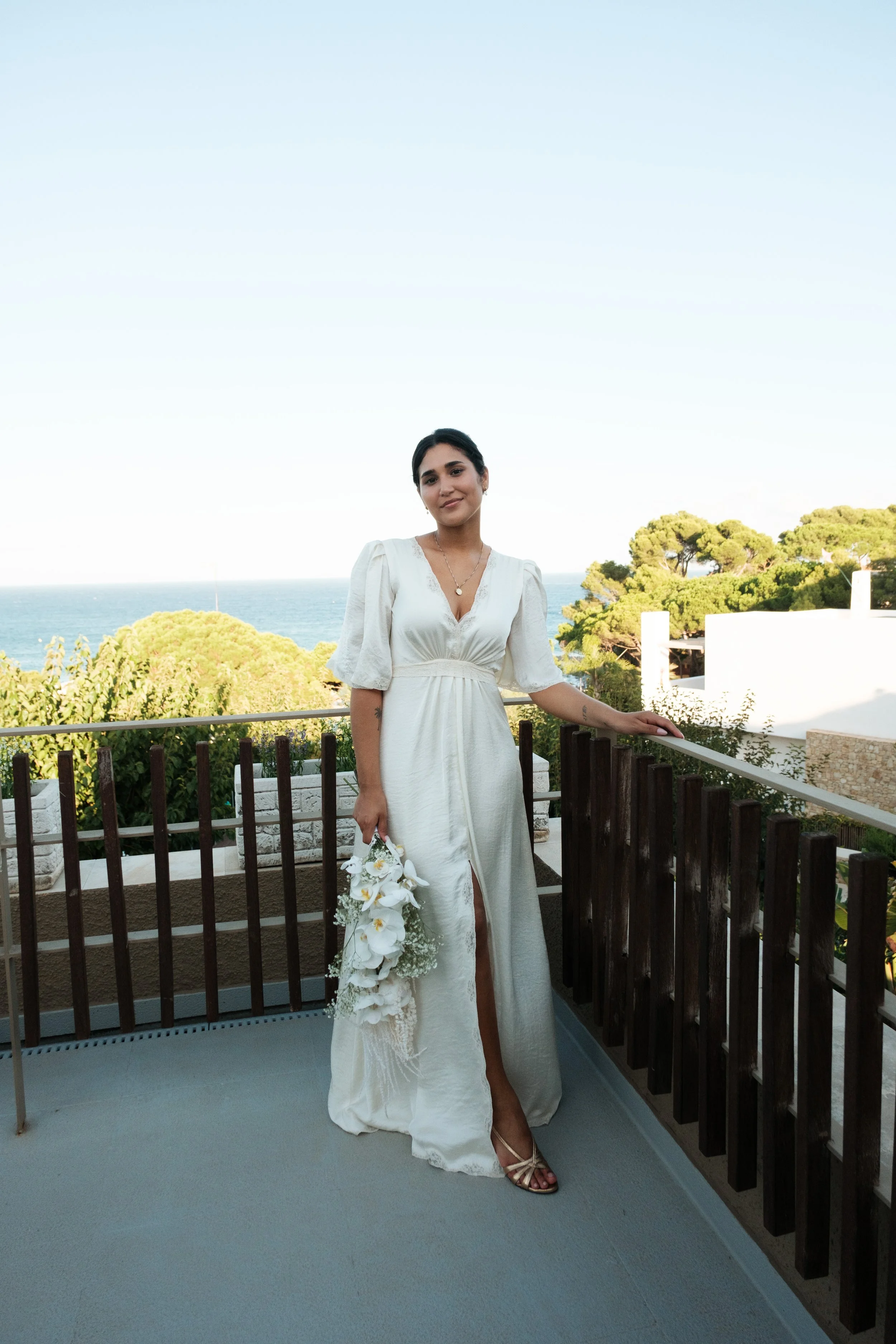 Eine junge Frau in einem weißen Brautkleid steht auf einem Balkon, hält einen Blumenstrauß und blickt lächelnd in die Kamera, mit Blick auf das Meer und grüne Bäume im Hintergrund.