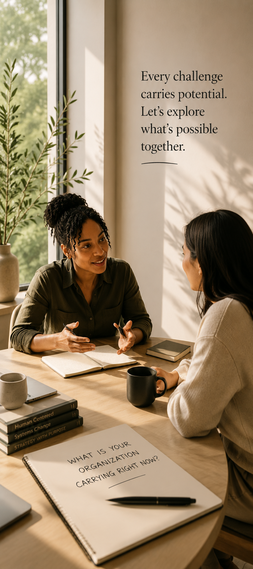 Two women are sitting at a desk engaged in a conversation in a well-lit room with sunlight streaming through a large window. On the desk are notebooks, a laptop, mugs, and a notepad with the question, "What is your organization carrying right now?" The background wall has an inspirational quote, "Every challenge carries potential. Let’s explore what’s possible together."