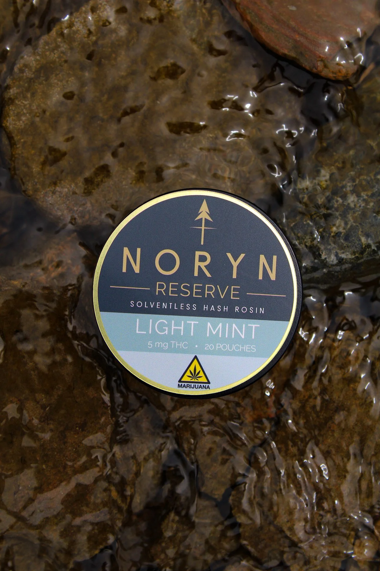 A package of NORYN Reserve solventless hash rosin with light mint flavor, placed on wet rocks in water.