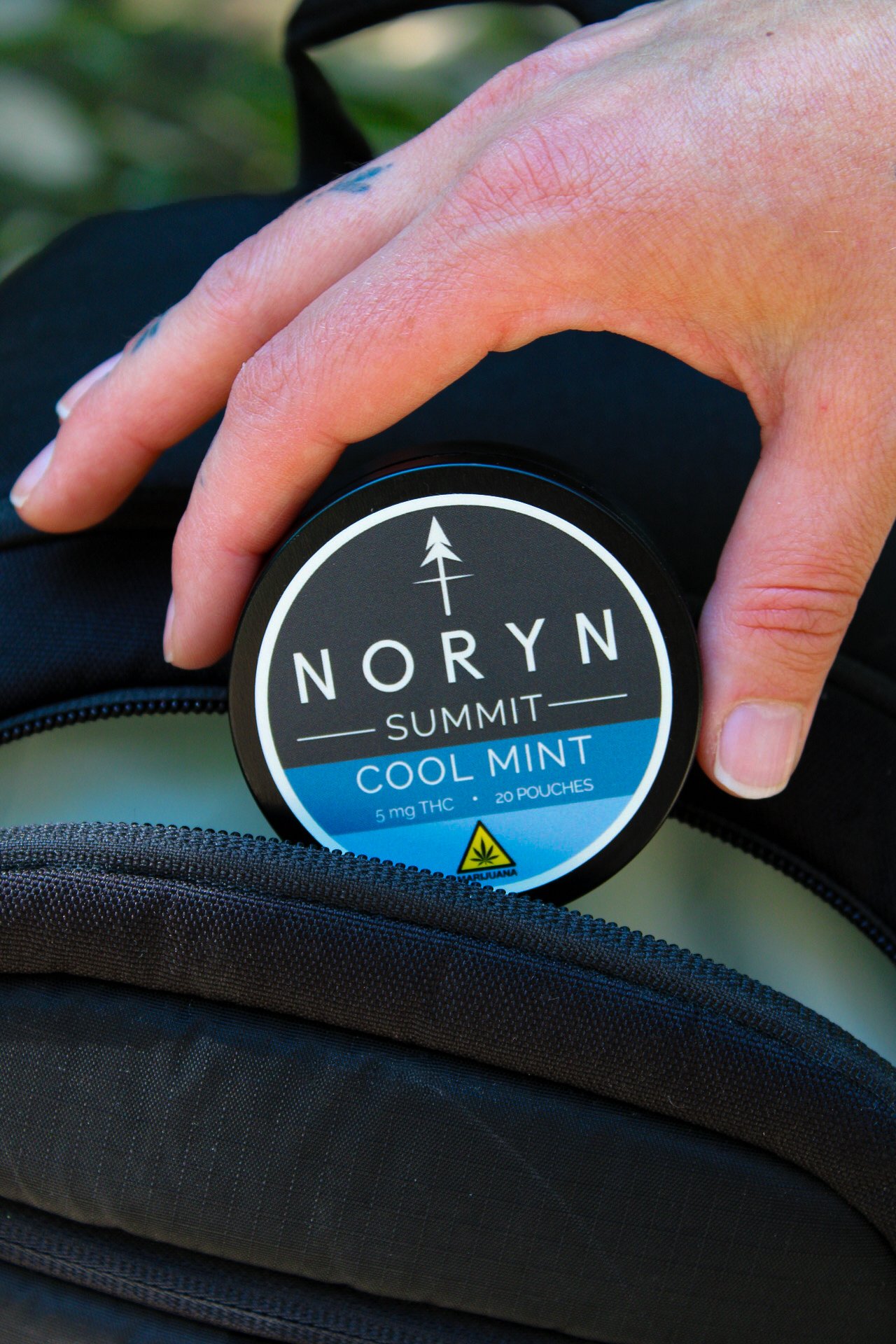 Close-up of a person holding a container of NORYN Summit Cool Mint THC pouches, with a focus on their hand and the product label, in front of a black backpack.