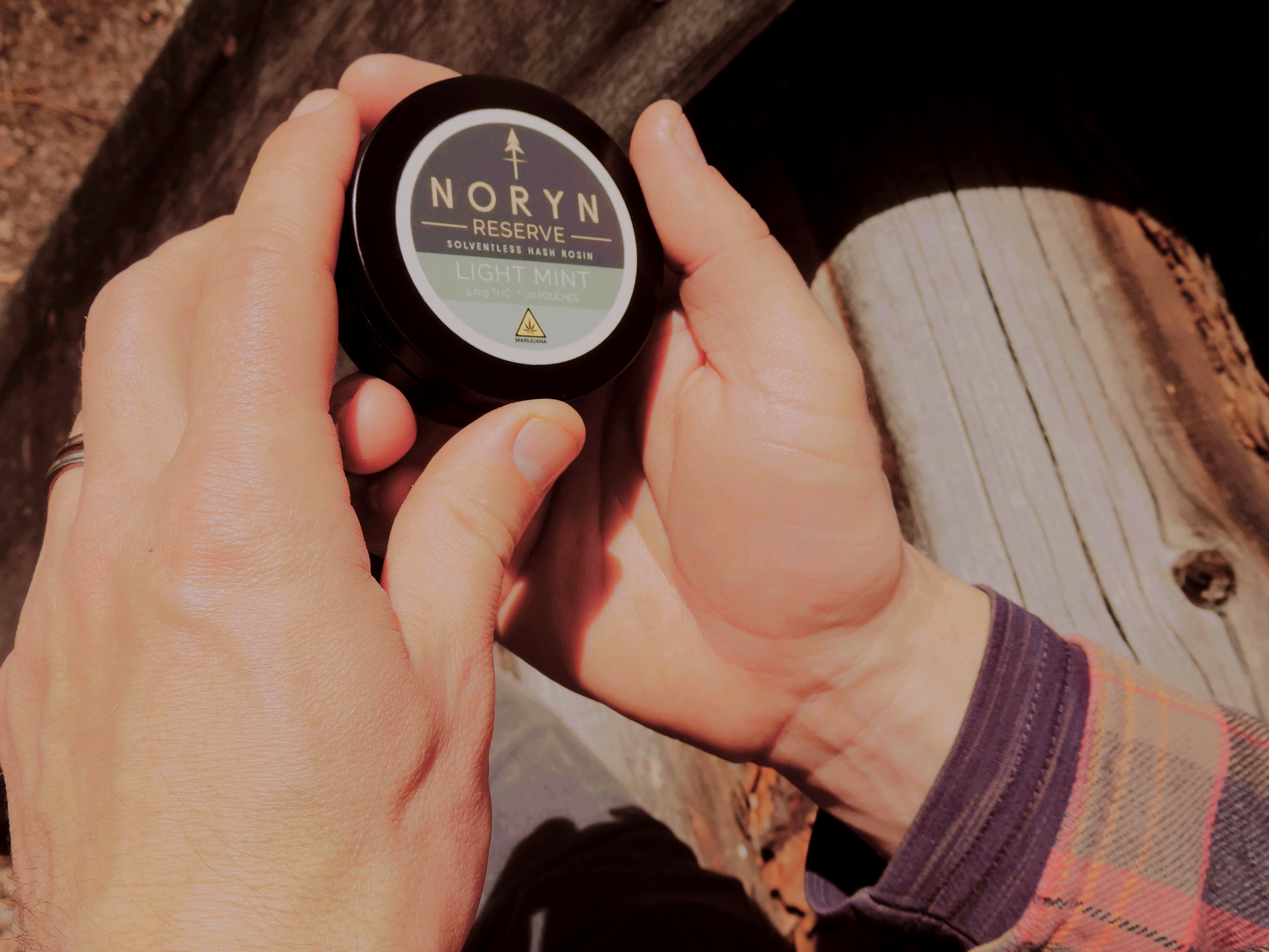 Person holding a black container labeled 'Noryn Reserve' with a description of THC content and flavor 'Light Mint'.