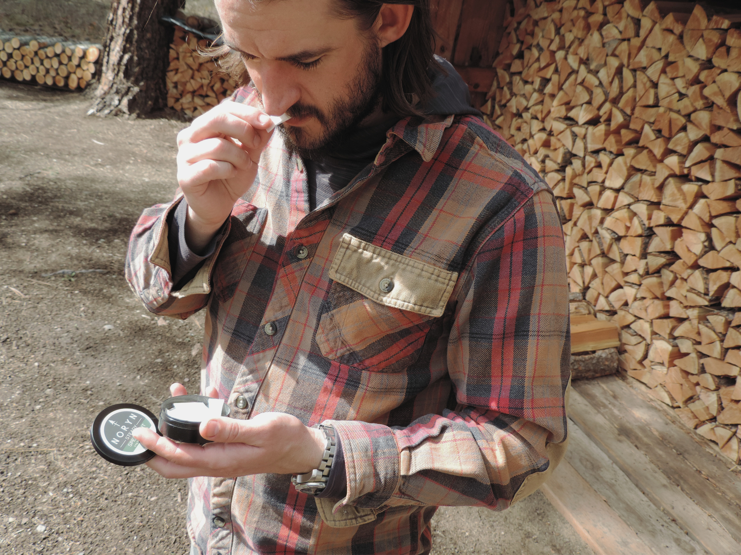 A man in a plaid shirt is holding a small round container and smelling a white substance, standing outdoors near a woodpile.