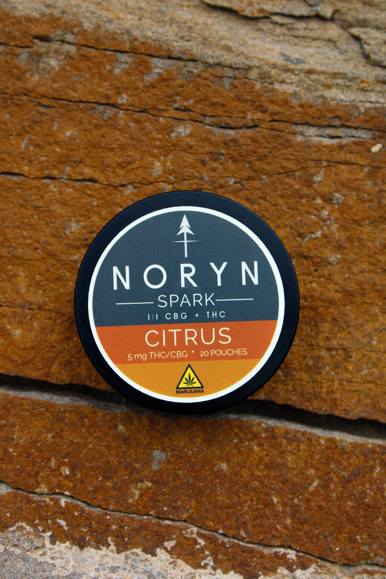 Close-up of a circular container label with the text "NORYN Spark Citrus" and information about THC and CBG content, placed on a textured orange-brown rock surface.