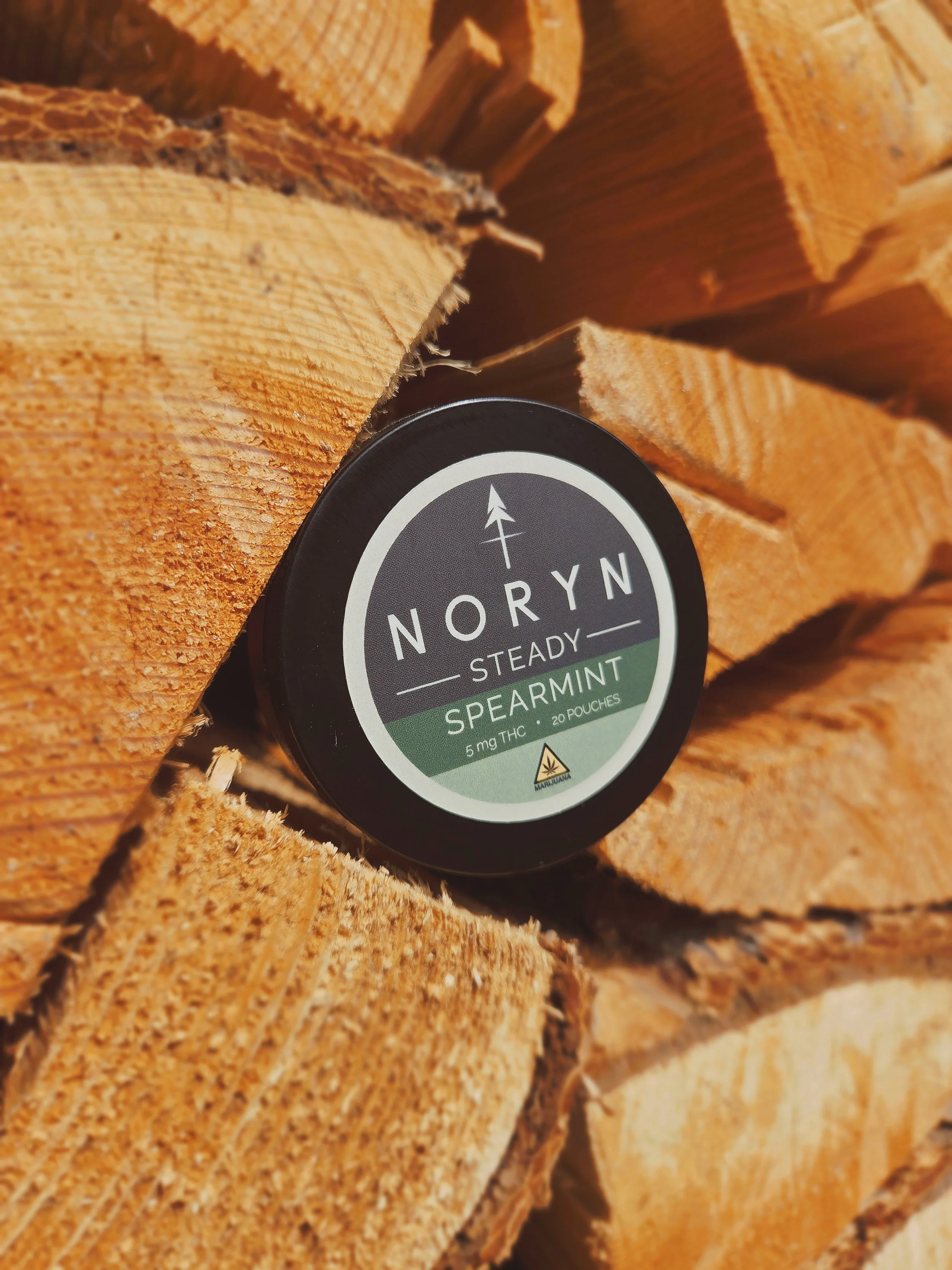 A black container labeled 'NORYN Steady Spearmint' with 5 mg THC, placed against a background of stacked firewood outdoor.