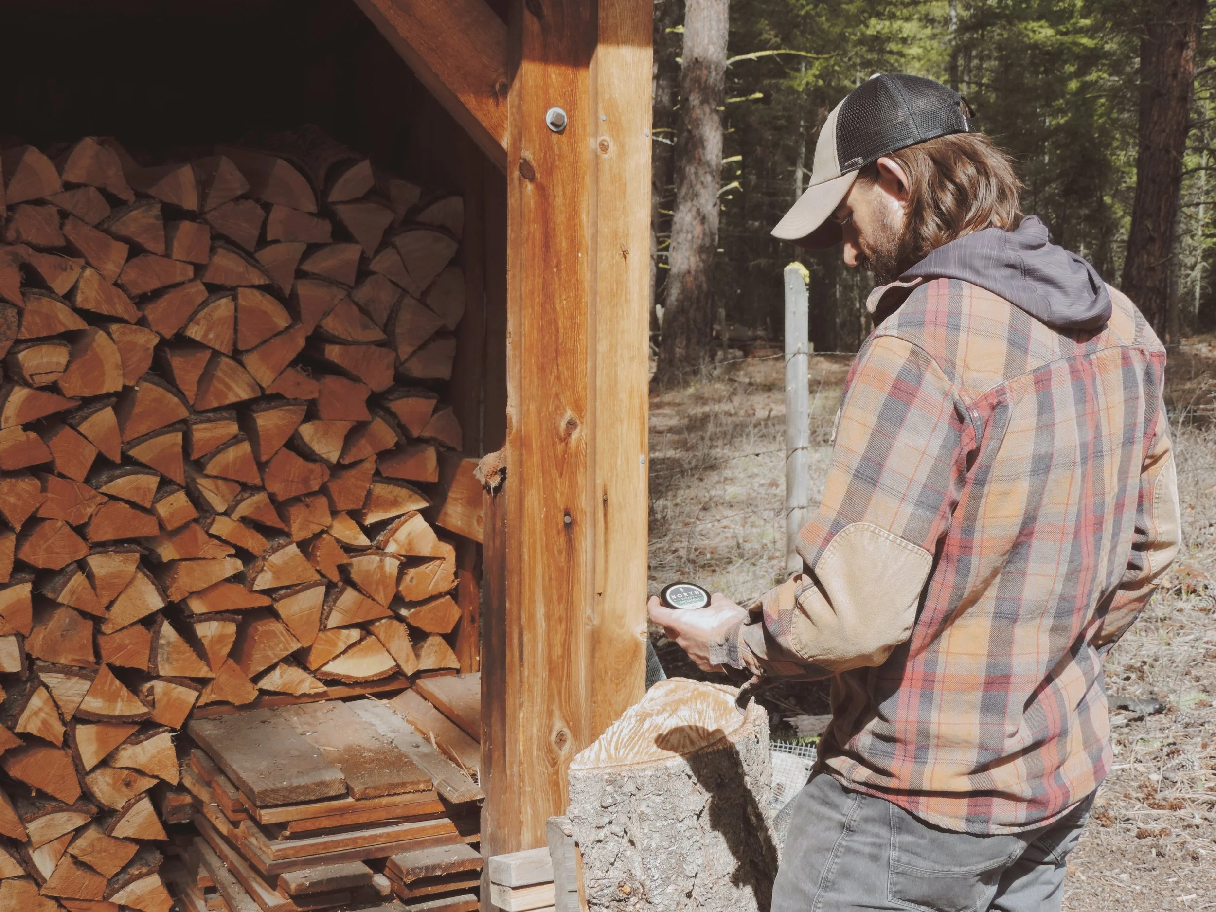 Man in plaid shirt and baseball cap looking at a pocket compass outside near stacked firewood.