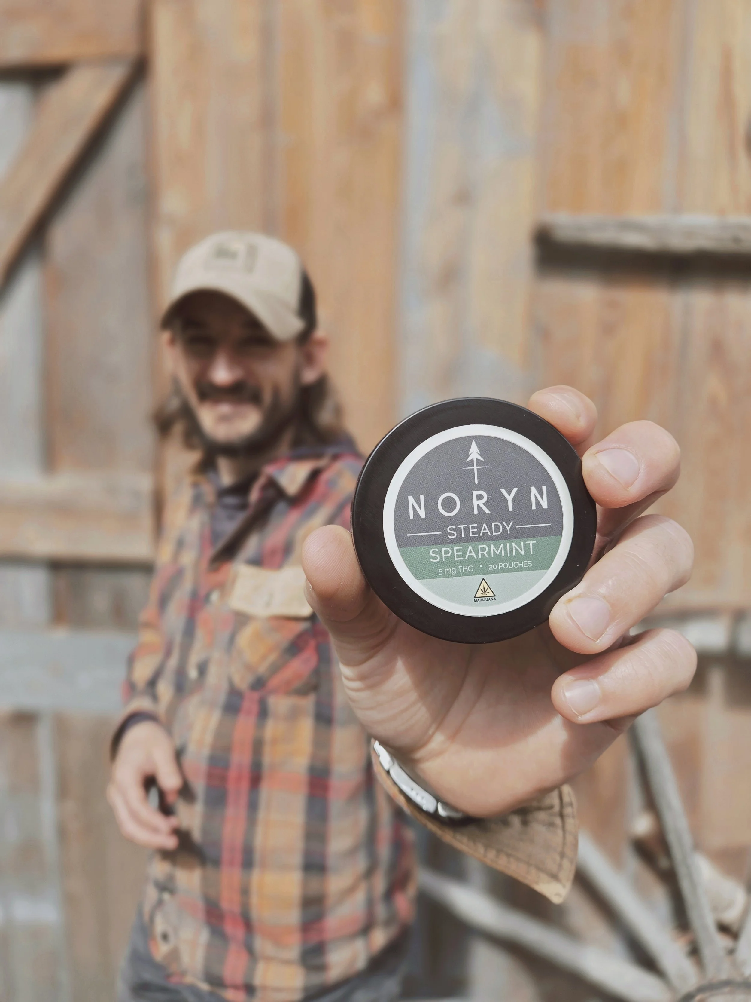 Man holding a container of Noryn Spearmint cannabis product with a wooden barn in the background.