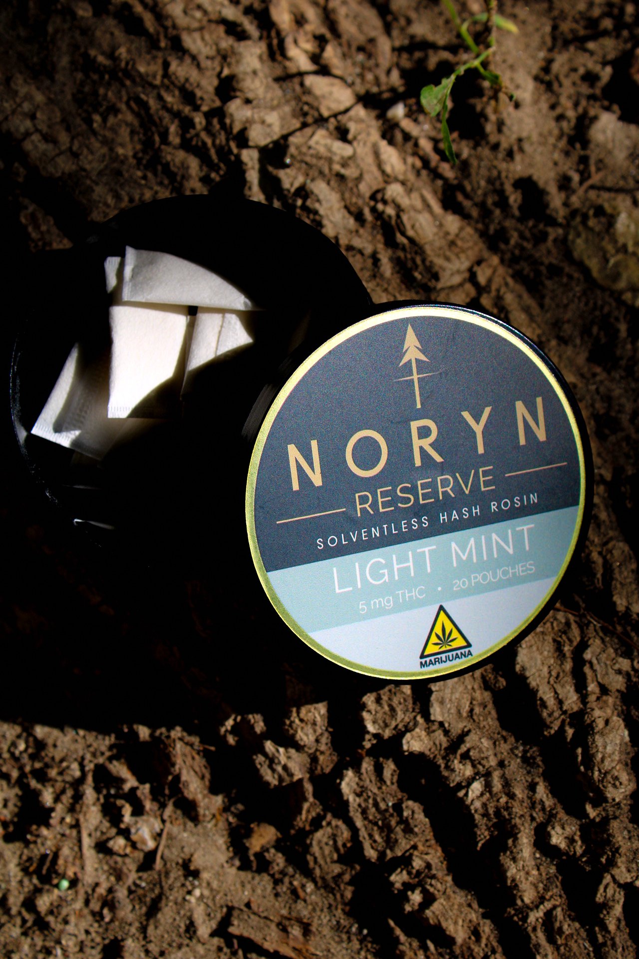 A container of NORYN Reserve Light Mint cannabis product lying on a tree, with the lid partially open revealing white pouches inside.