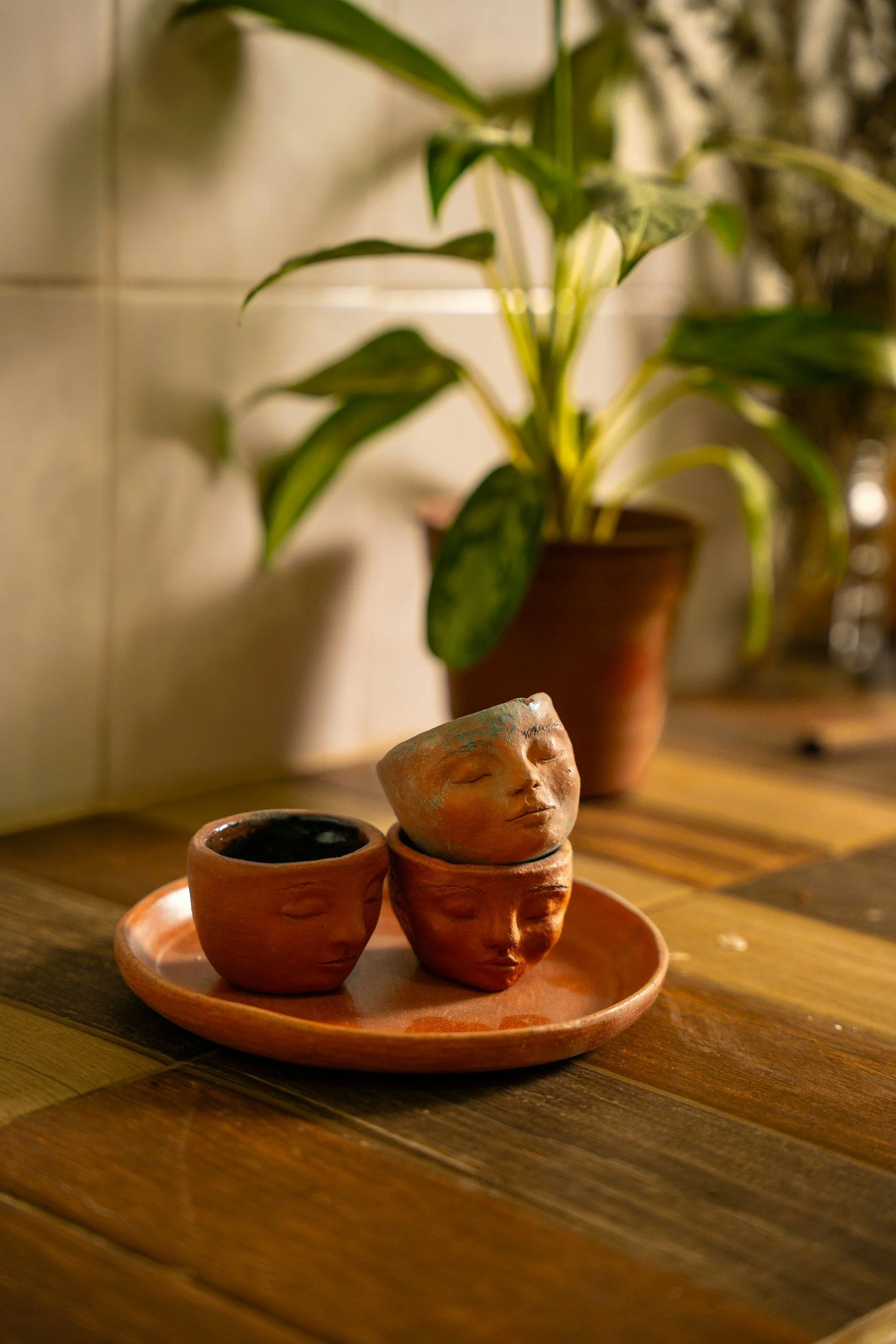 Decorative clay cups with human faces arranged on a circular tray on a wooden surface, with a potted plant in the background.