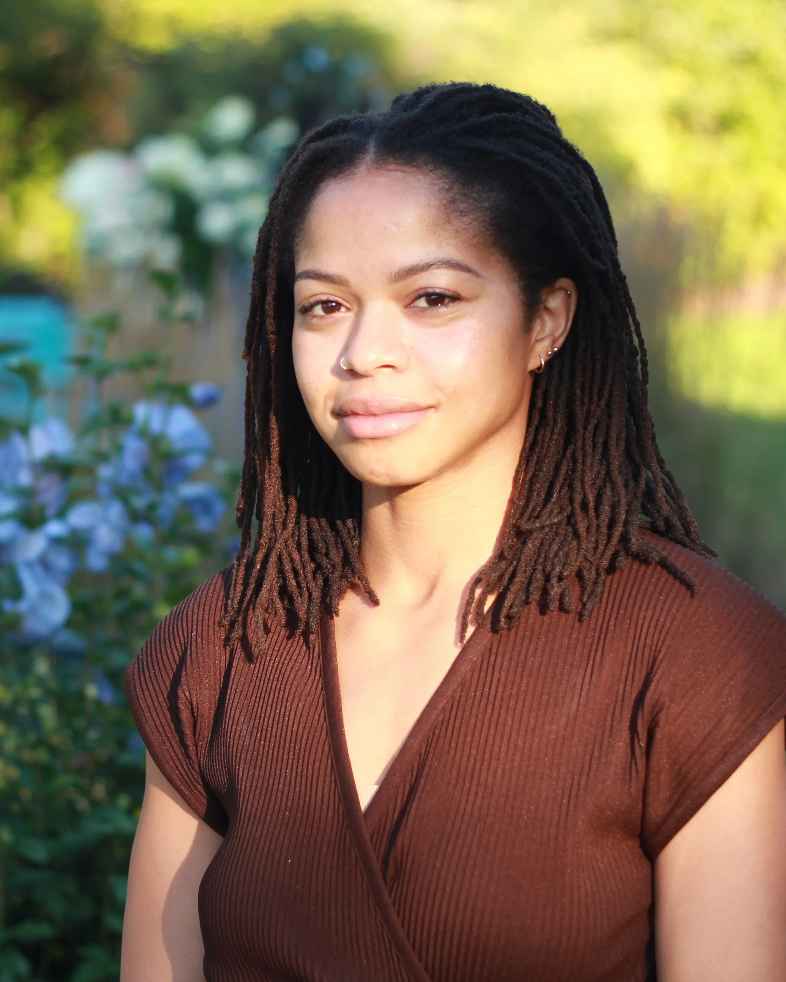 A young woman with dreadlocks and a nose piercing, wearing a brown top, standing outdoors in front of greenery and flowers.