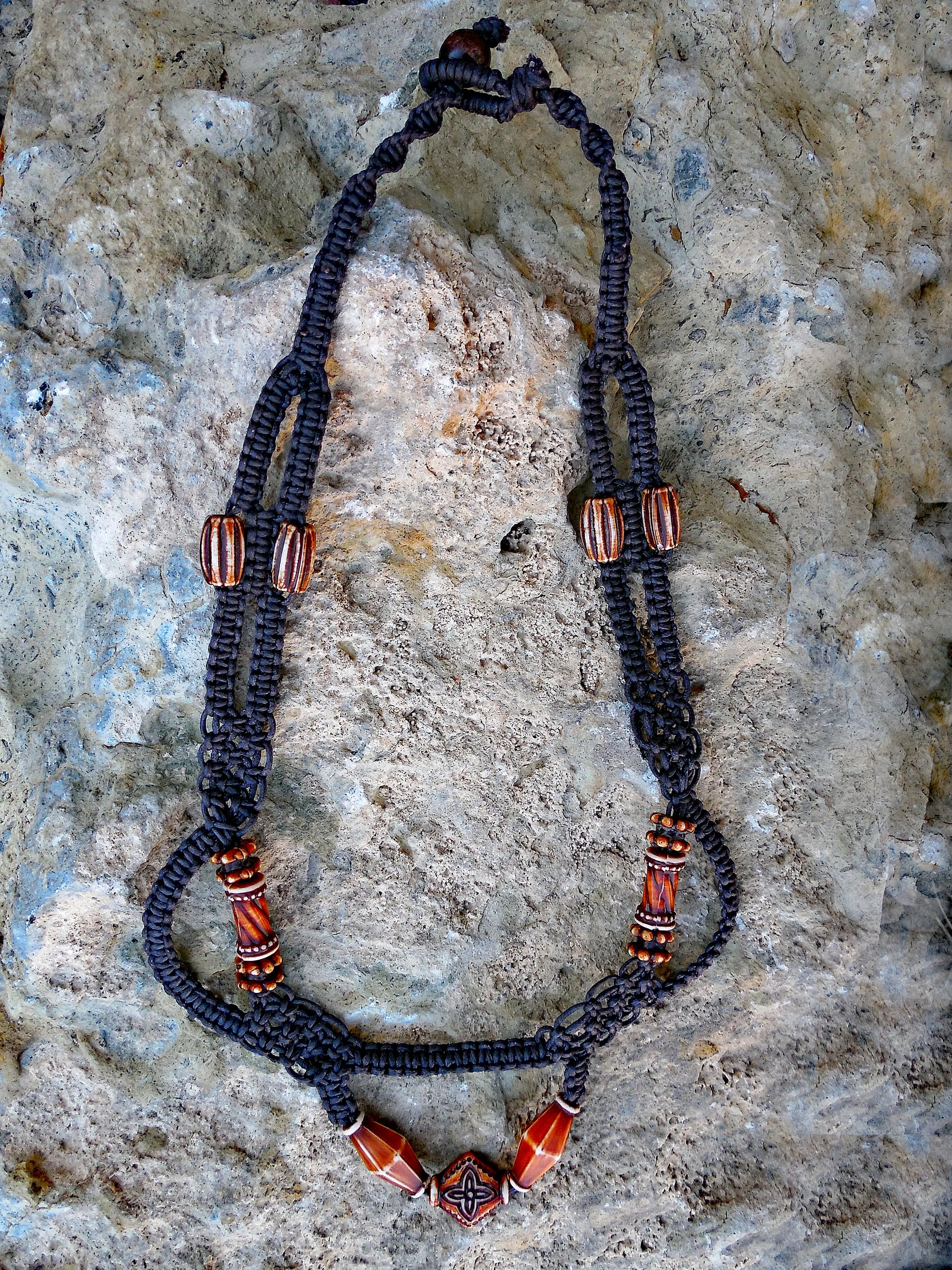 A necklace with black braided cord and large orange and black patterned beads, laid on a rocky surface.