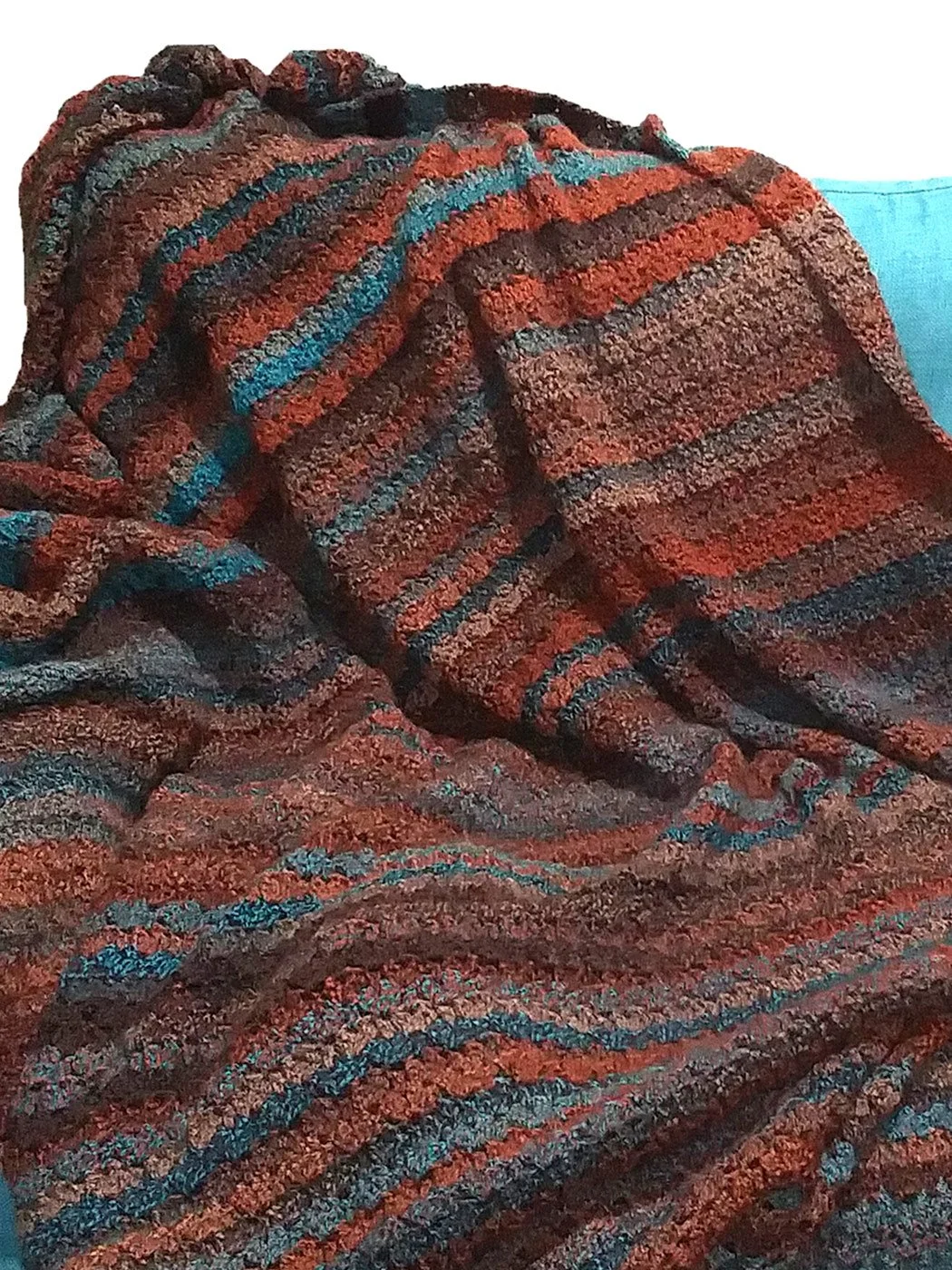 An oversized crochet blanket in rust, brown, and teal tones draped cozy style for the Painted Desert Thick Blanket pattern featured in the October 2026 newsletter.