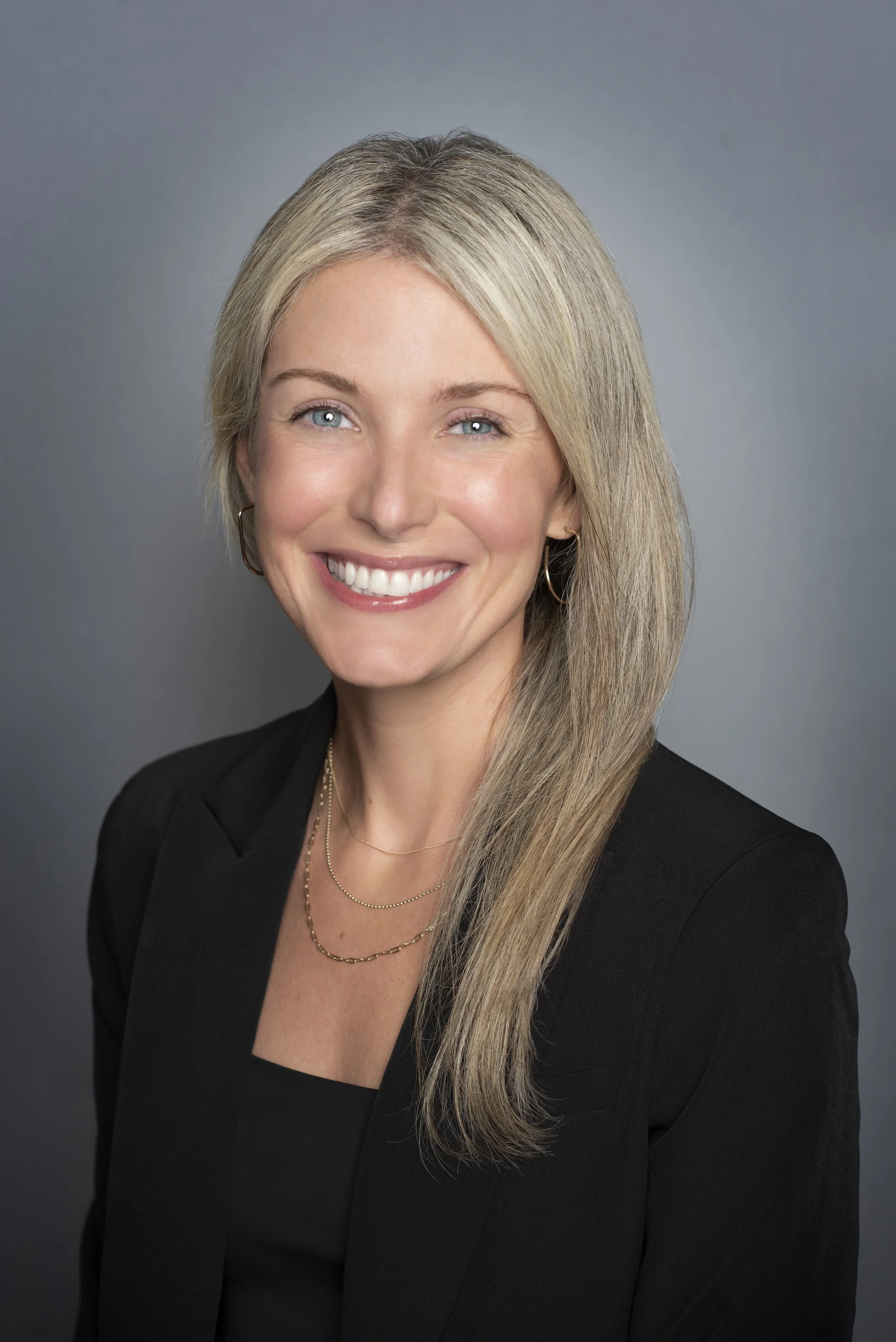 A professional woman with long blond hair, blue eyes, and a bright smile, wearing a black blazer and layered necklaces, standing against a gray background.