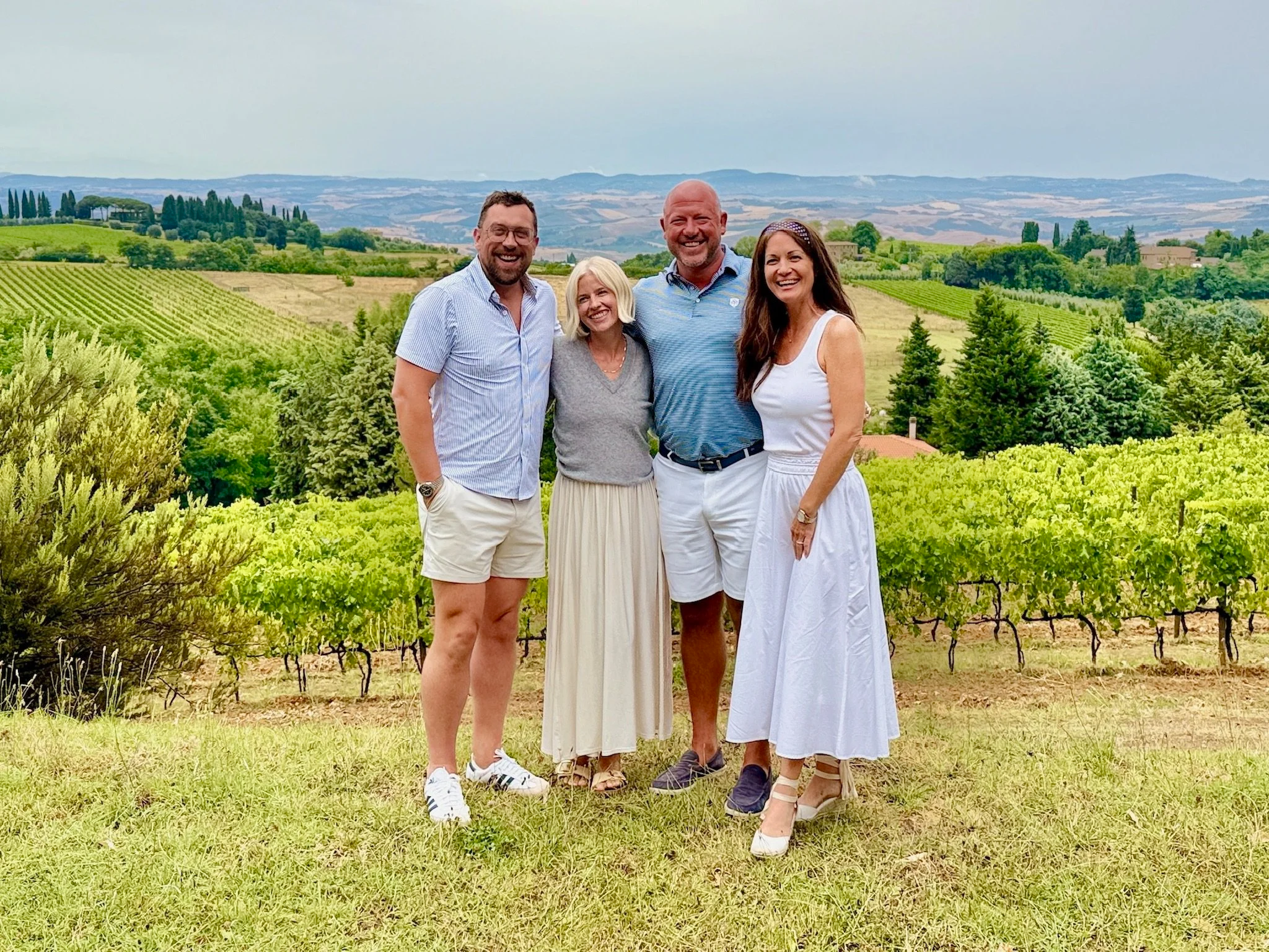 Group of four smiling people standing together in a vineyard with rows of grapevines and rolling hills in the background.
