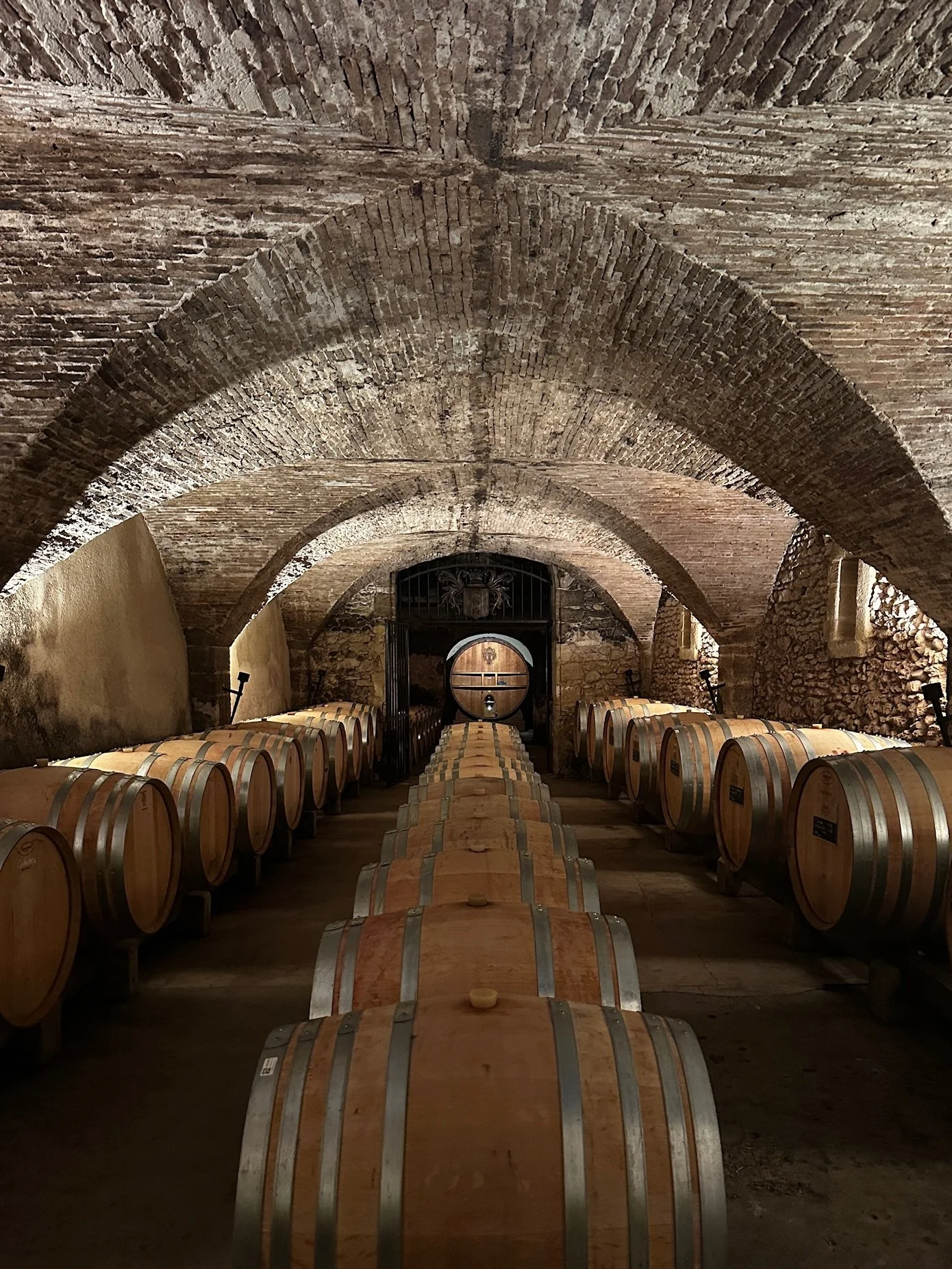 A wine cellar with a curved brick ceiling, stone walls, and wooden barrels lined up in rows.