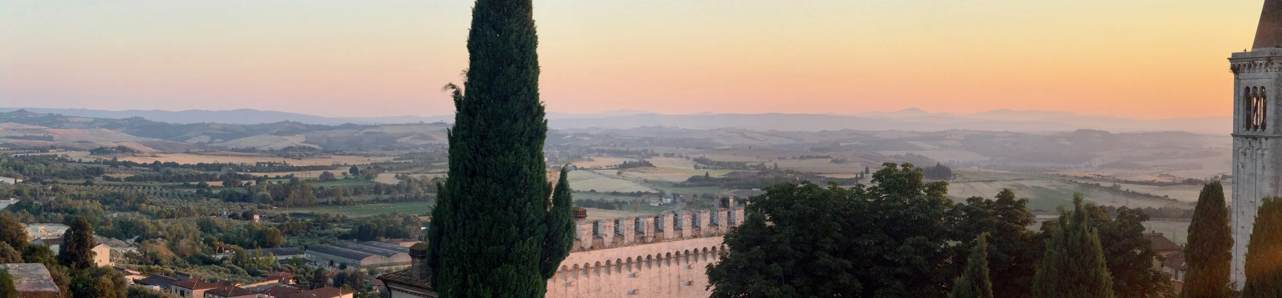 Panoramic view of Tuscan countryside at sunset, with rolling hills, cypress trees, and historic stone buildings, including a tower on the right.