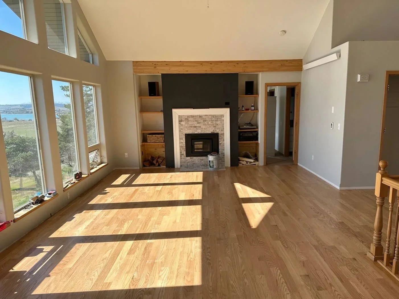 Empty living room with large windows, hardwood floors, a fireplace with a brick surround, built-in shelves, and a view of a lake outside.