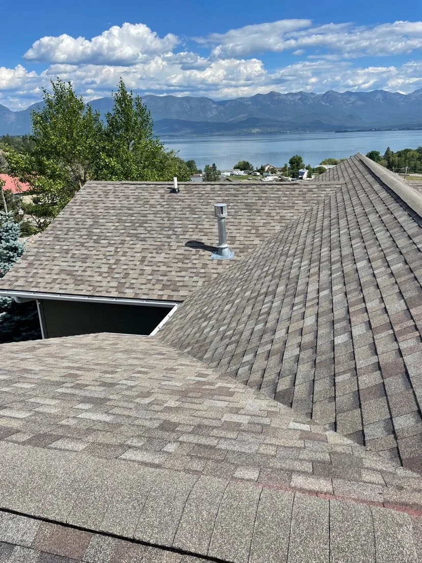 View of rooftops with shingles, trees, a body of water, and mountains in the background under a partly cloudy sky.