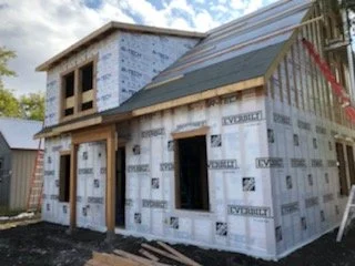 A two-story house under construction with unfinished exterior walls and windows, and scaffolding nearby.