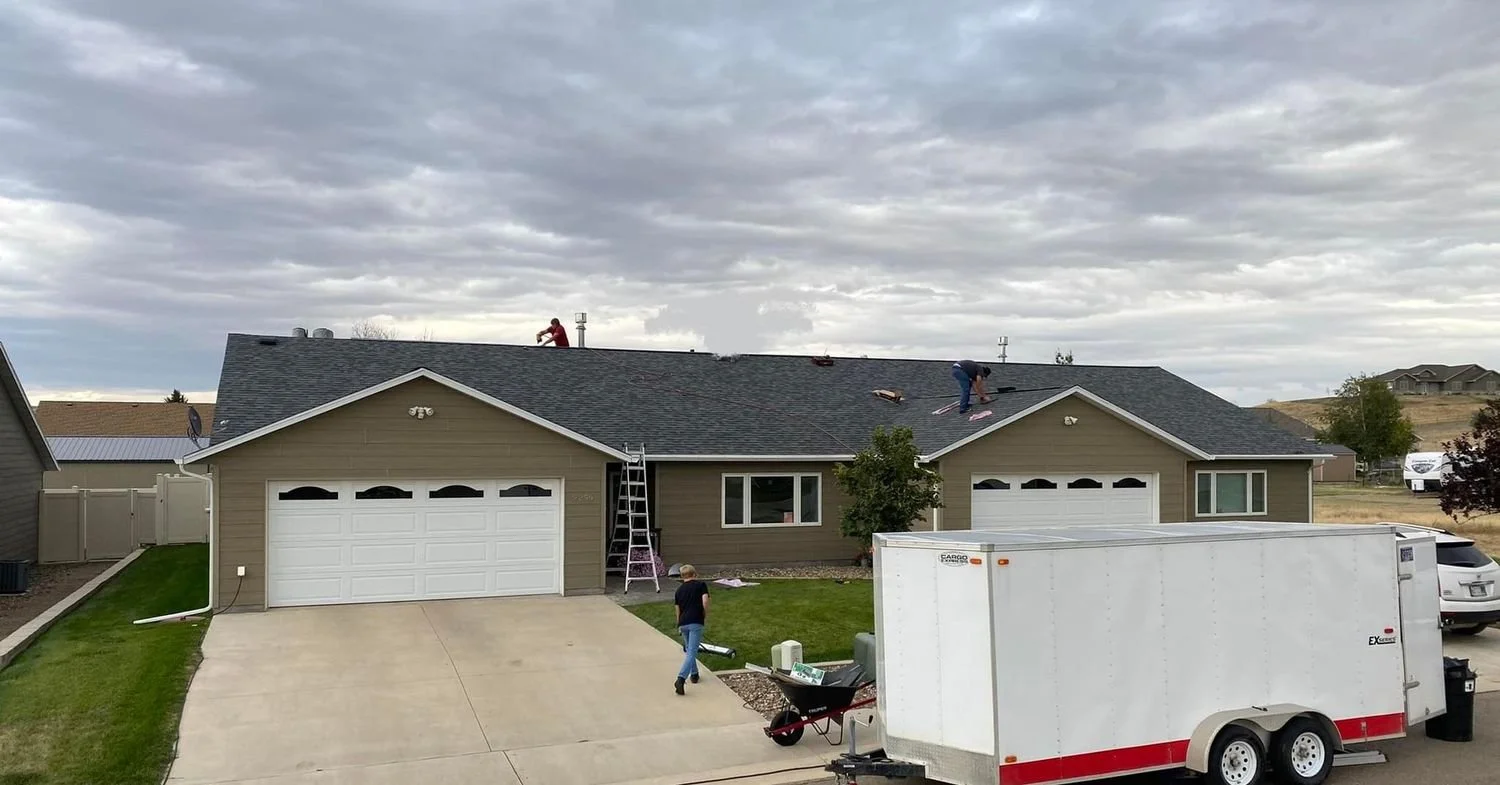 Two workers on a house roof installing or repairing shingles, with a ladder leaning against the house. A person is walking in the driveway while a large white trailer is parked nearby. The house has a two-car garage, and the sky is cloudy.