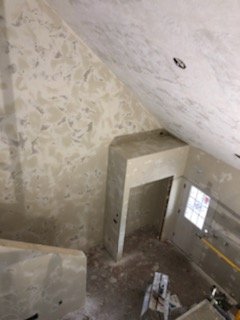 View of an attic space with a sloped ceiling, unfinished drywall, a small window, and construction materials on the floor.