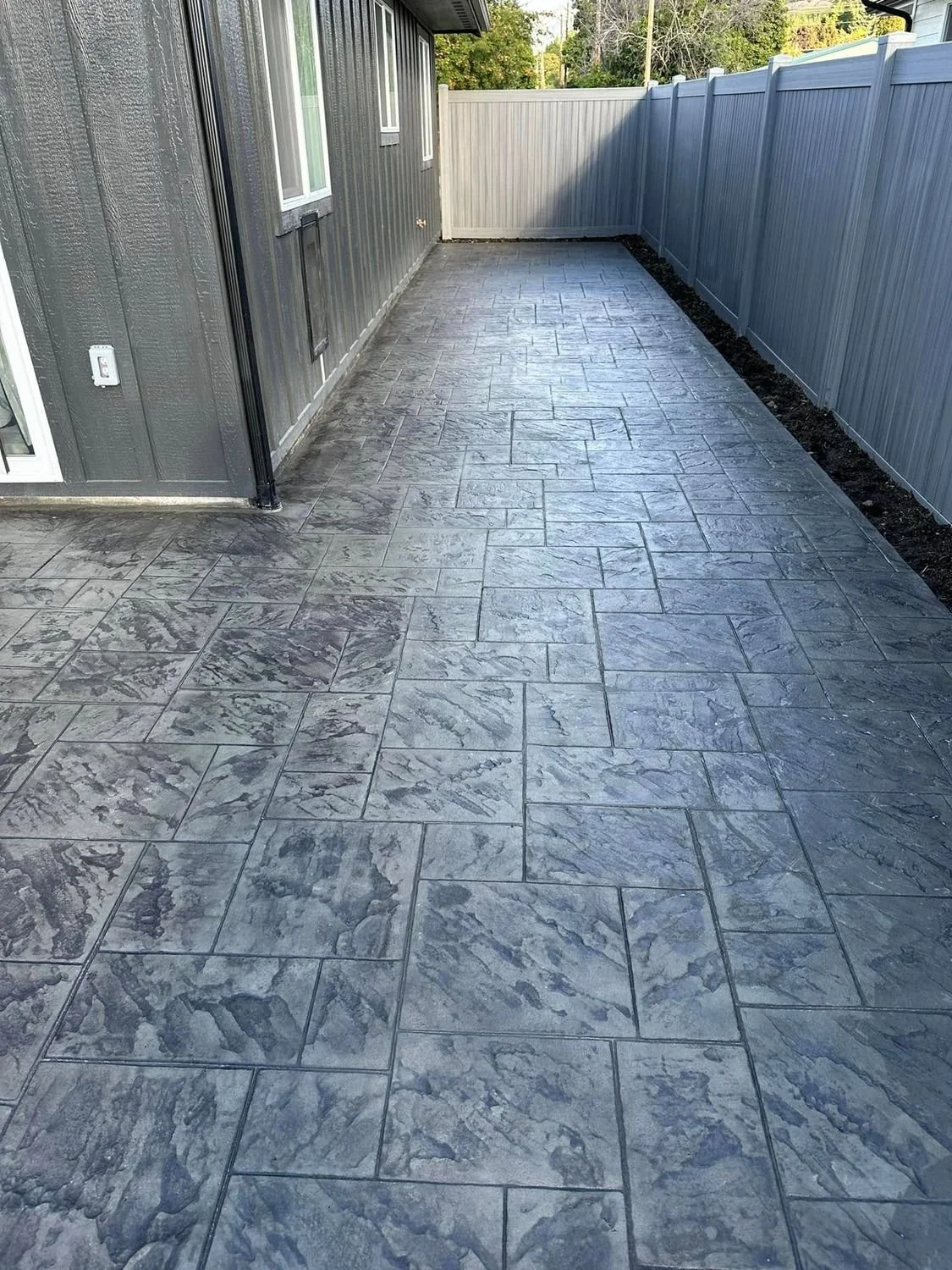 Backyard patio with gray stamped concrete, black siding house on the left, gray fencing on the right, and mulch along the fencing.