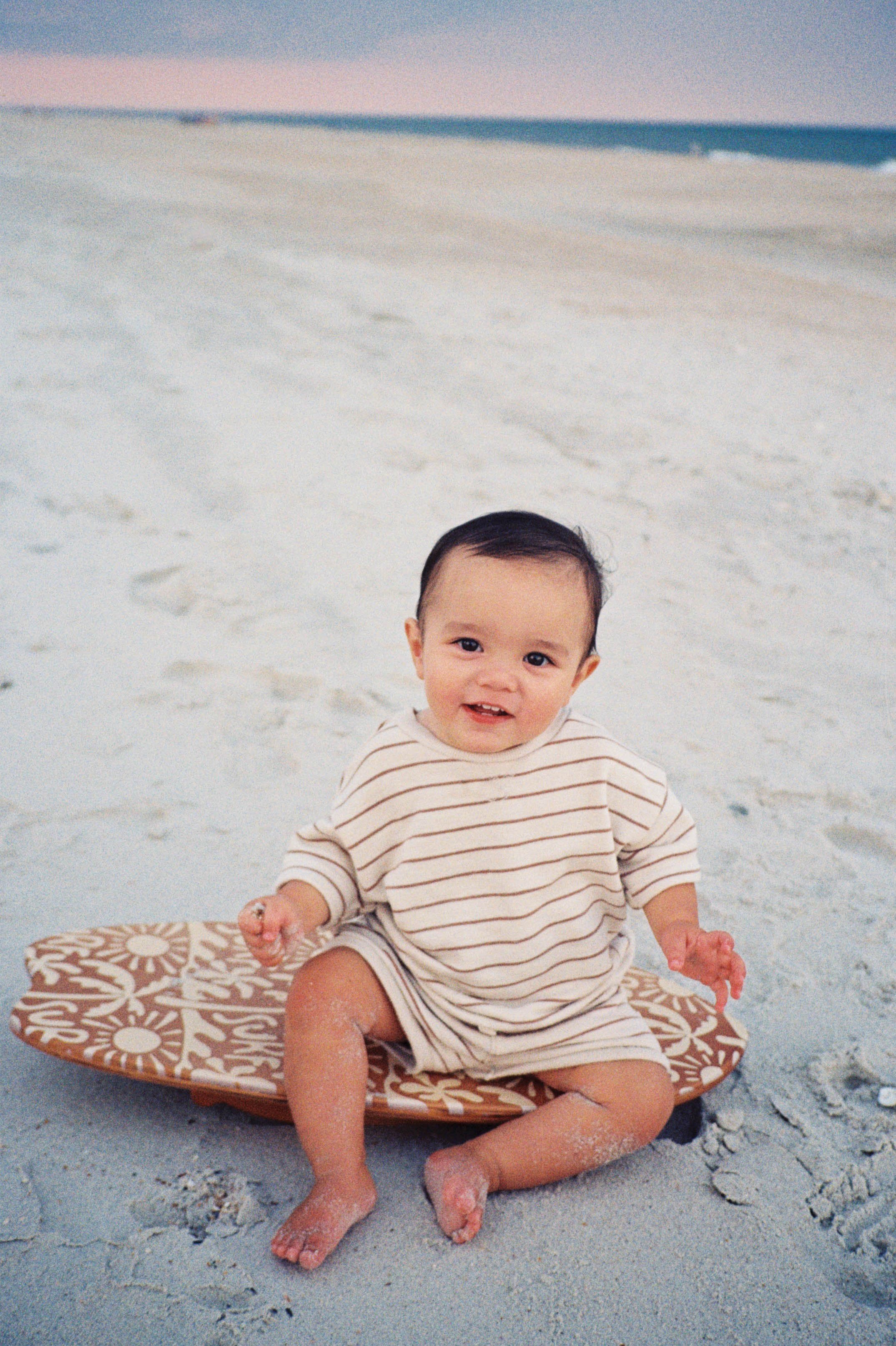Happy baby sitting on a surfboard at the beach, embodying the coastal spirit behind Swell Smiles Pediatric Dentistry in Jacksonville