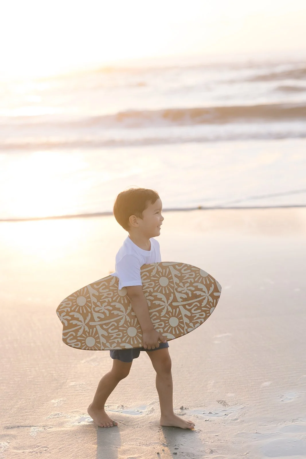 Young boy sitting on the beach at golden hour with a surfboard, representing childhood in Jacksonville, FL at Swell Smiles