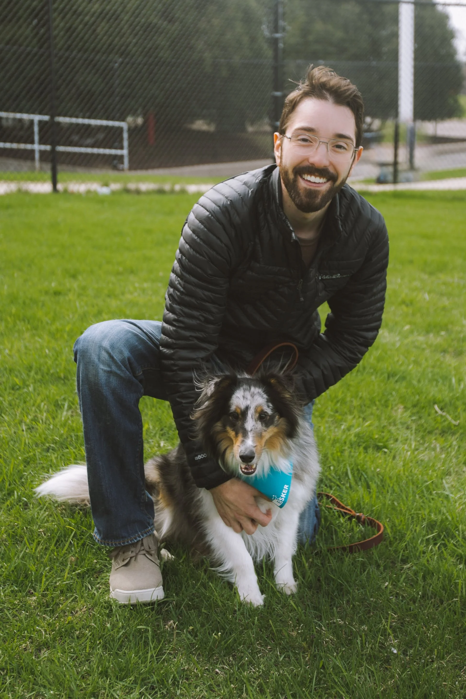A man with glasses and a beard smiling, kneeling on the grass, holding a dog with a blue bandana, in a park with green grass and a sports field fence in the background.