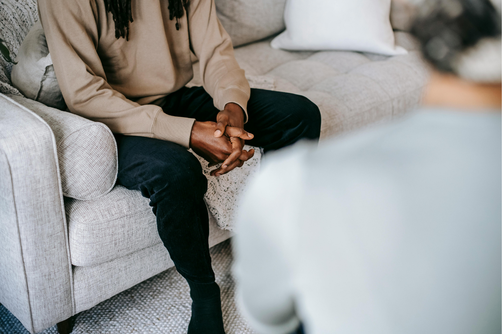 A person sitting on a light-colored sofa with black pants, a beige sweatshirt, and hair with dreadlocks, while another person, whose face is out of focus, is in the foreground, possibly engaged in conversation.