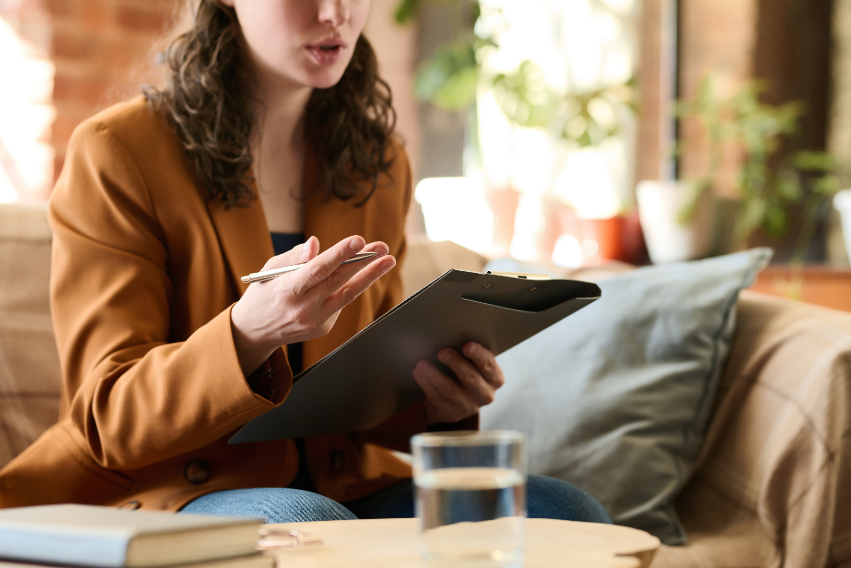 A woman sitting on a beige couch holding a clipboard and a pen, in a cozy room with natural light, books, and plants in the background.