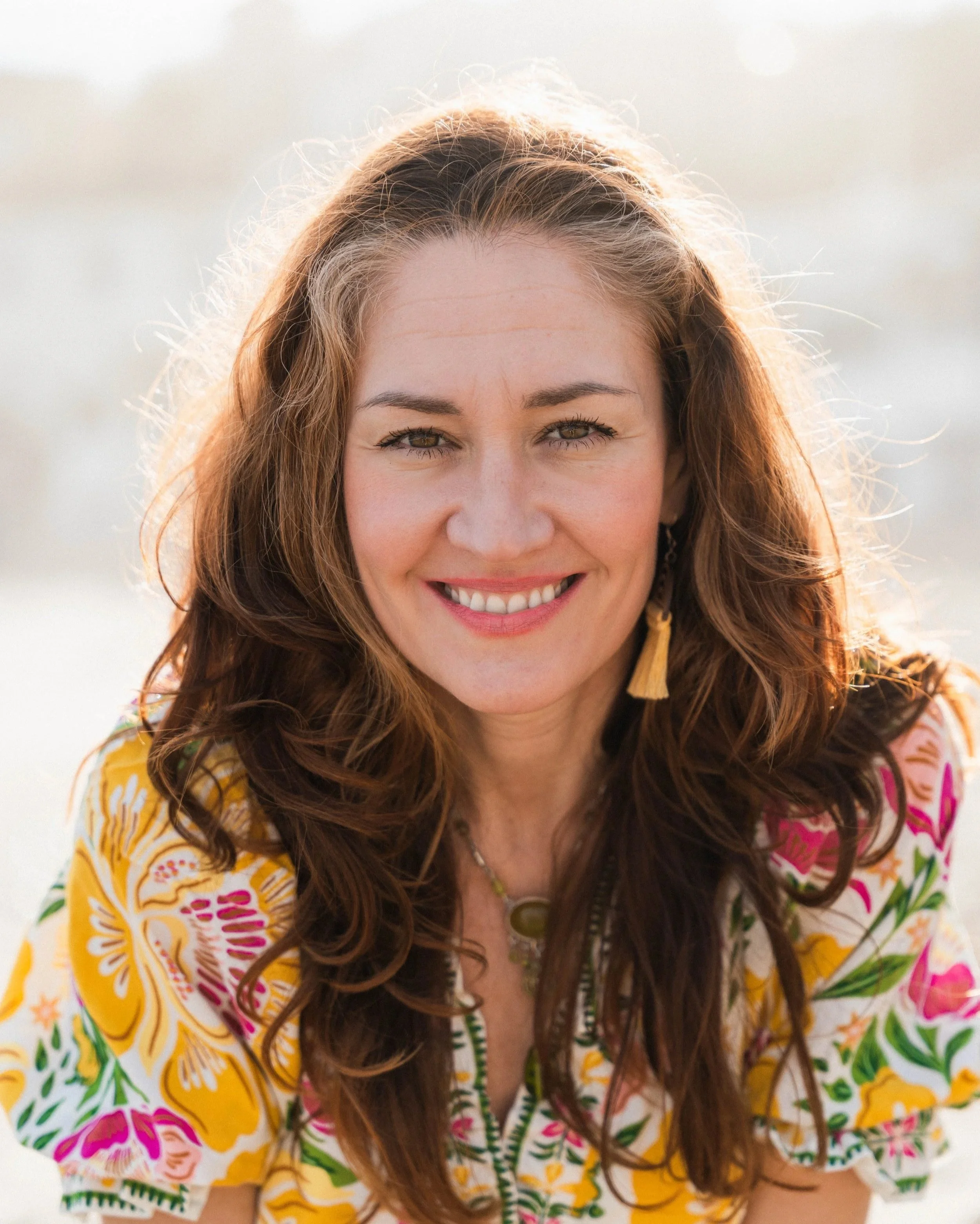 A woman with long curly brown hair smiling, wearing a colorful dress with floral patterns, outdoors in natural lighting.