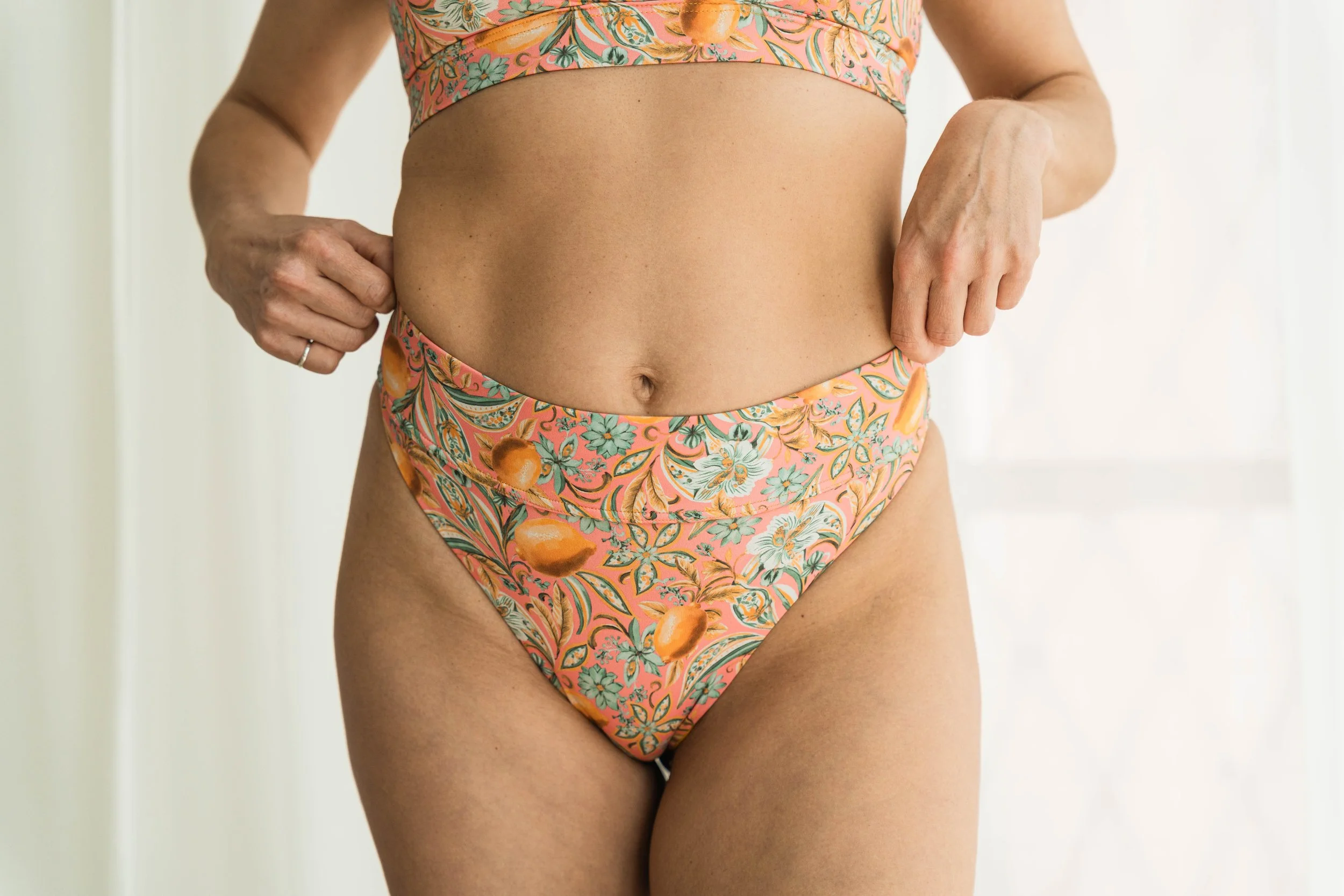 Close-up of a woman’s midsection wearing a floral bikini with peach, orange, and green colors. She is standing against a white background, with her hands resting on her hips.