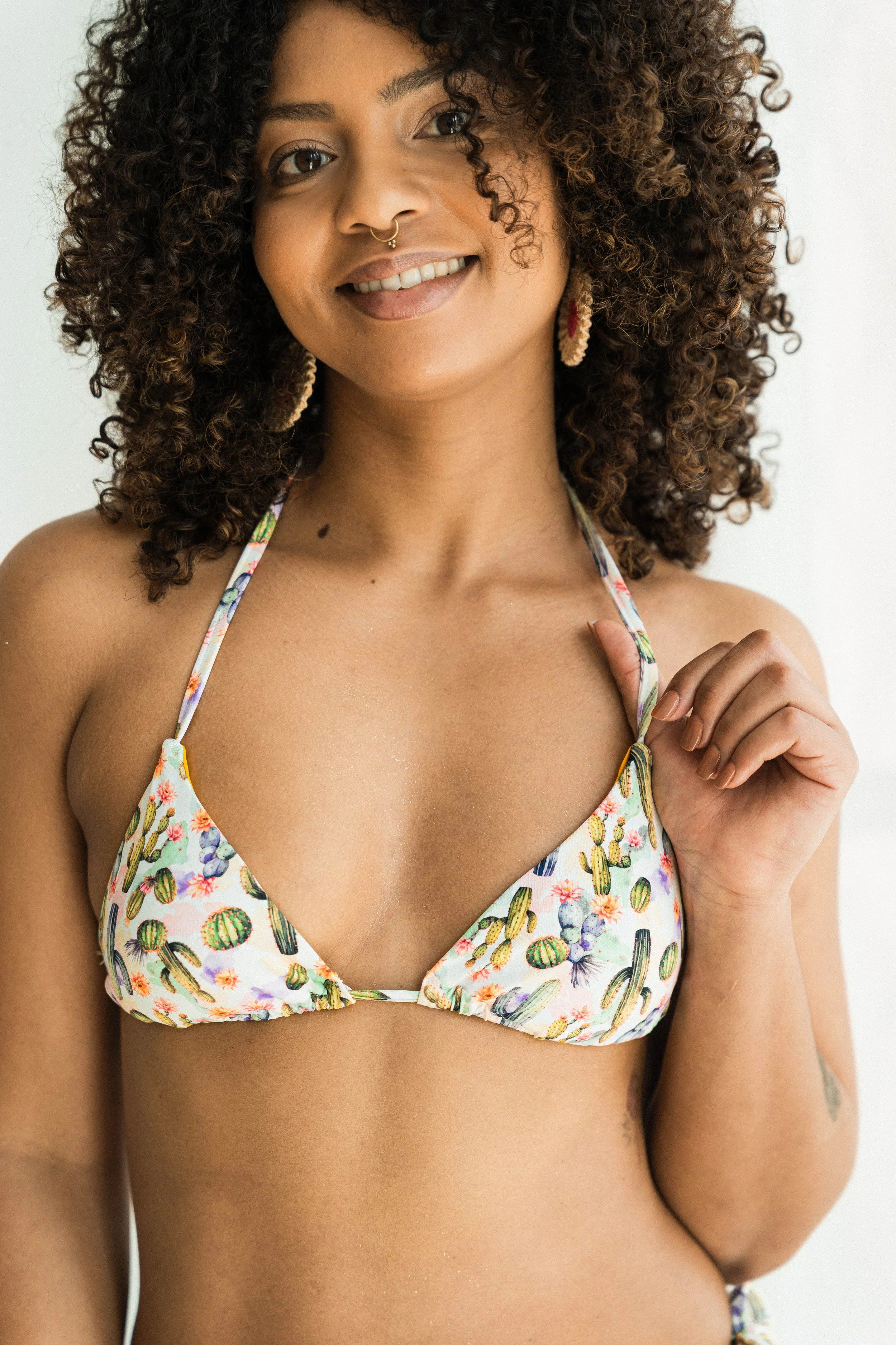 A woman with curly hair and tan skin is smiling, wearing a bikini top with a cactus and floral pattern, earrings, and a nose ring.