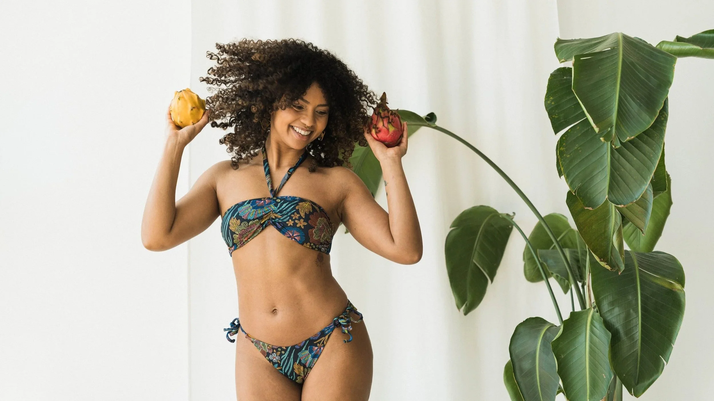 A woman with curly hair wearing a colorful bikini, holding a passion fruit and a dragon fruit, smiling, standing next to large green tropical leaves against a white background.
