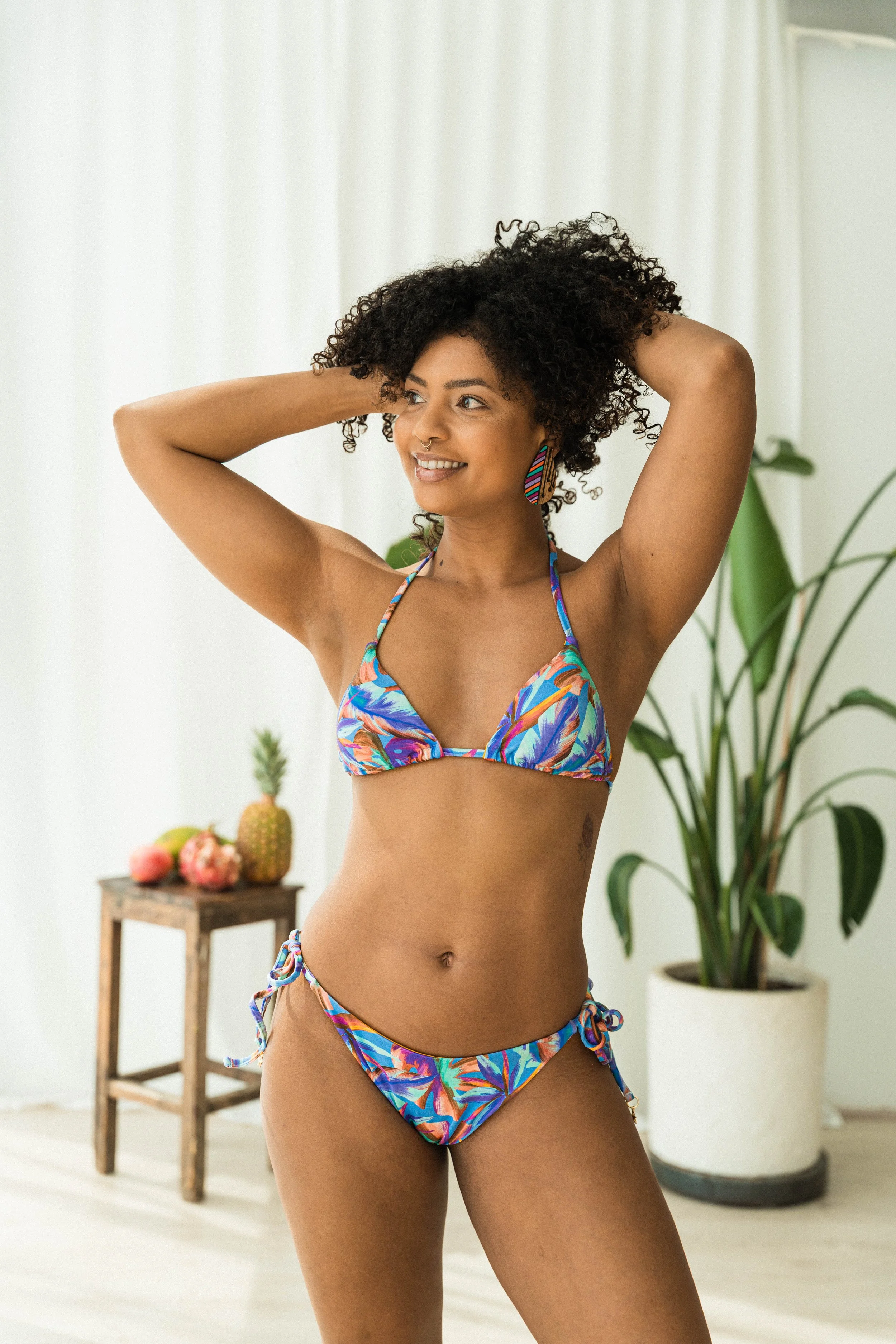 A woman in a colorful bikini with her hands behind her head, smiling in a bright room with a plant and a small wooden table with fruit in the background.