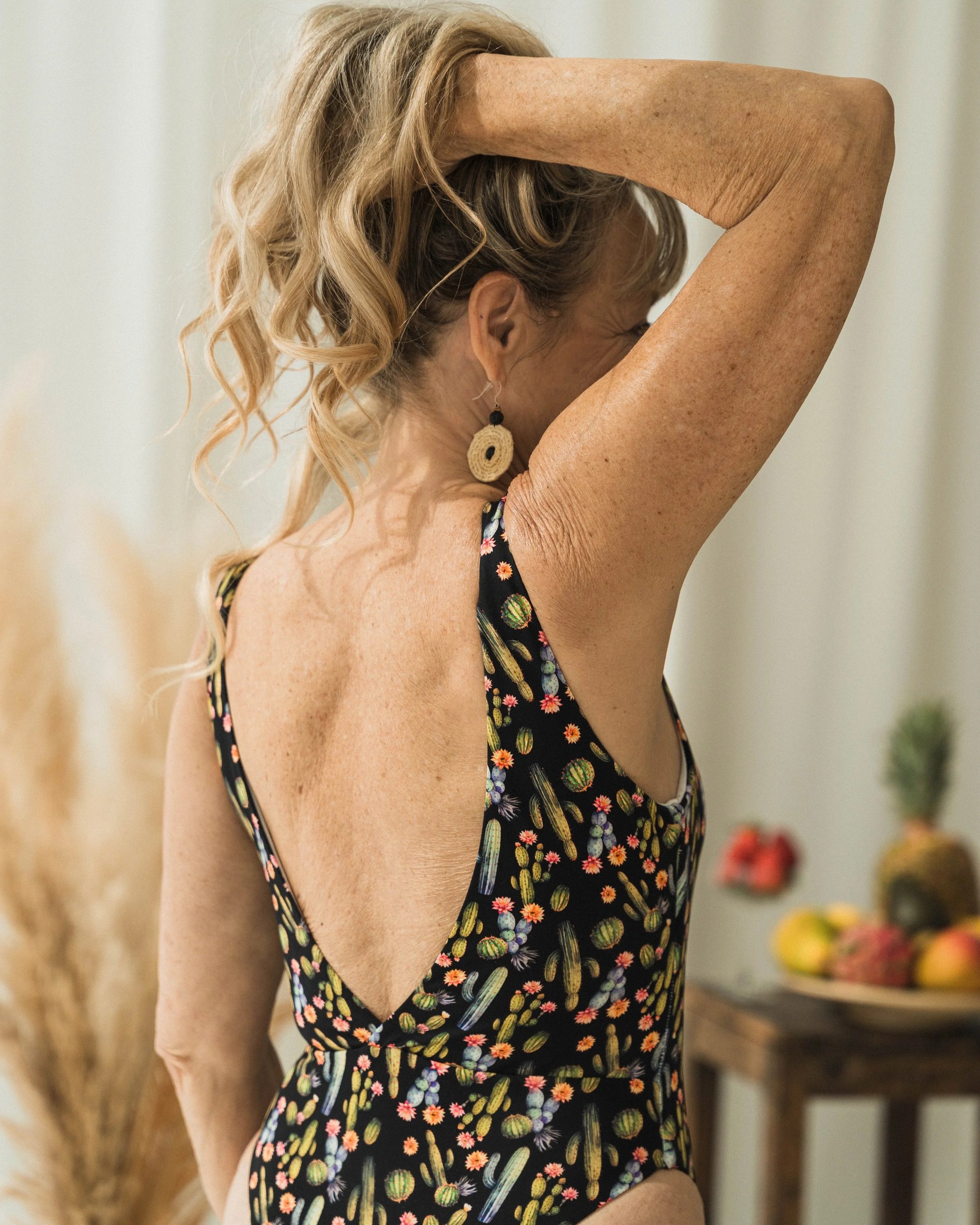 A woman with curly blonde hair wearing a black swimsuit with a colorful watermelon pattern, raising her left arm above her head, showing her shoulder and side profile in a room with light-colored walls and a shelf with decorative items in the background.