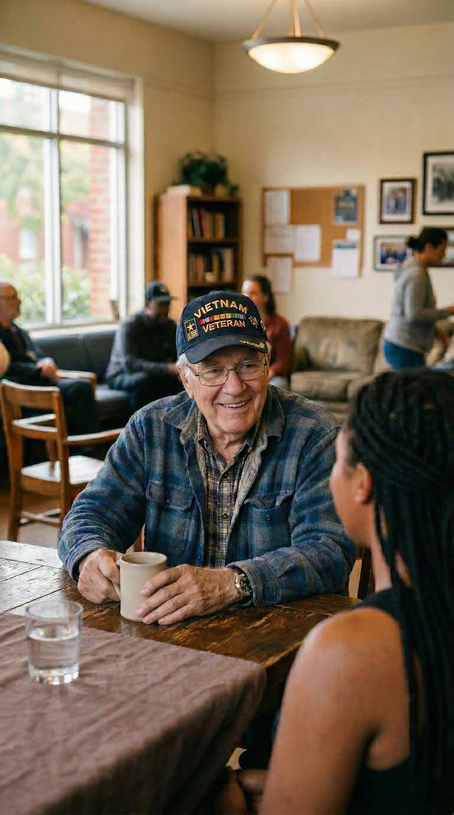 Older man wearing a Vietnam veteran hat and plaid shirt smiling while talking to a woman with dreadlocks in a cozy coffee shop or community center.