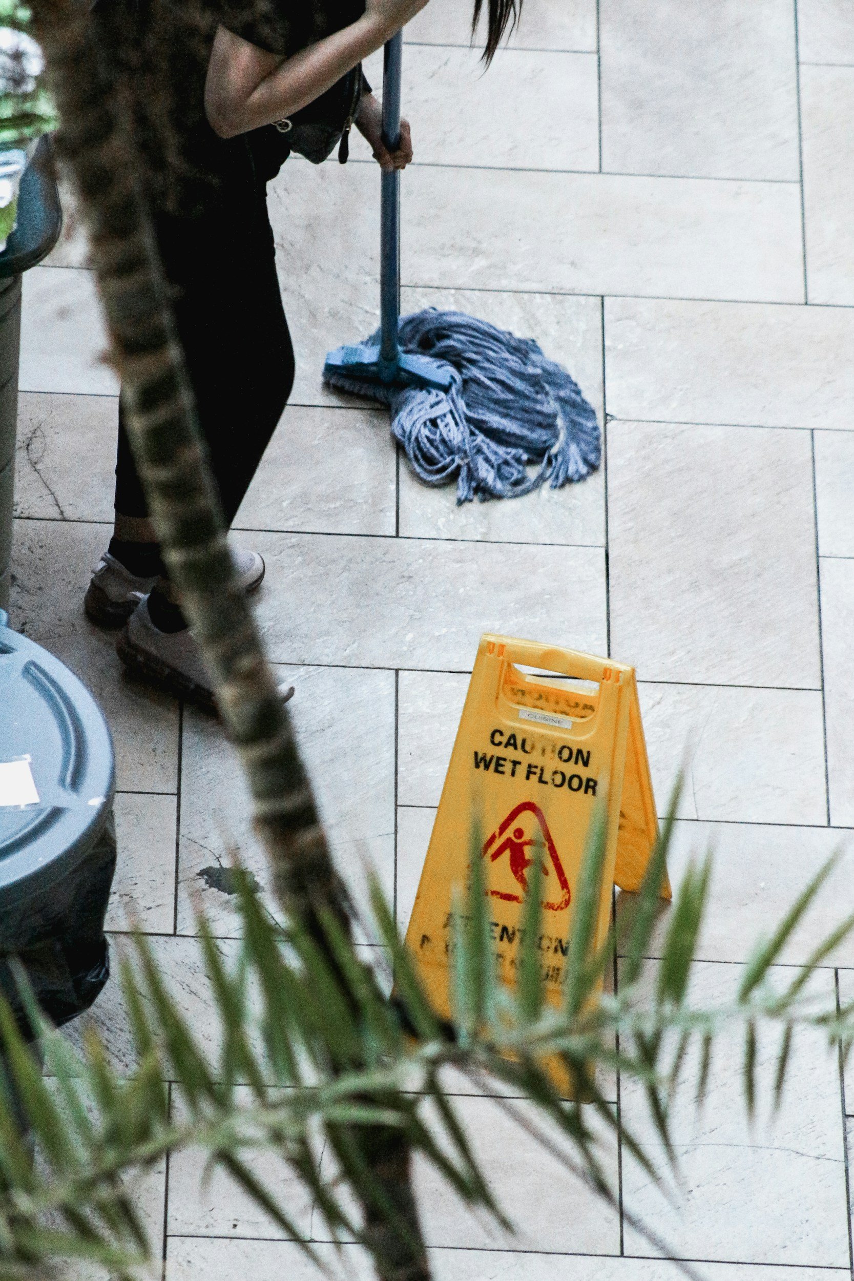 Mop resting on the floor next to a caution wet floor sign, with a person mopping the floor in the background.