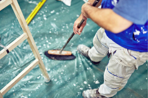 Person sweeping water on green plastic-covered floor beneath a wooden ladder.