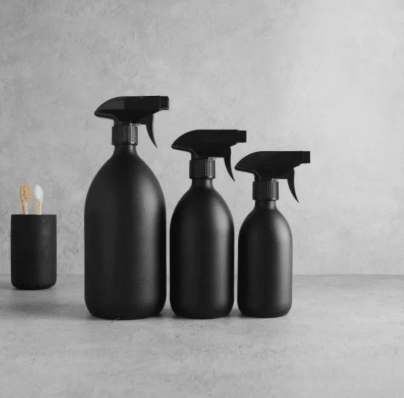 Three black spray bottles of varying sizes on a gray surface against a gray wall, with a small container holding cotton swabs nearby.