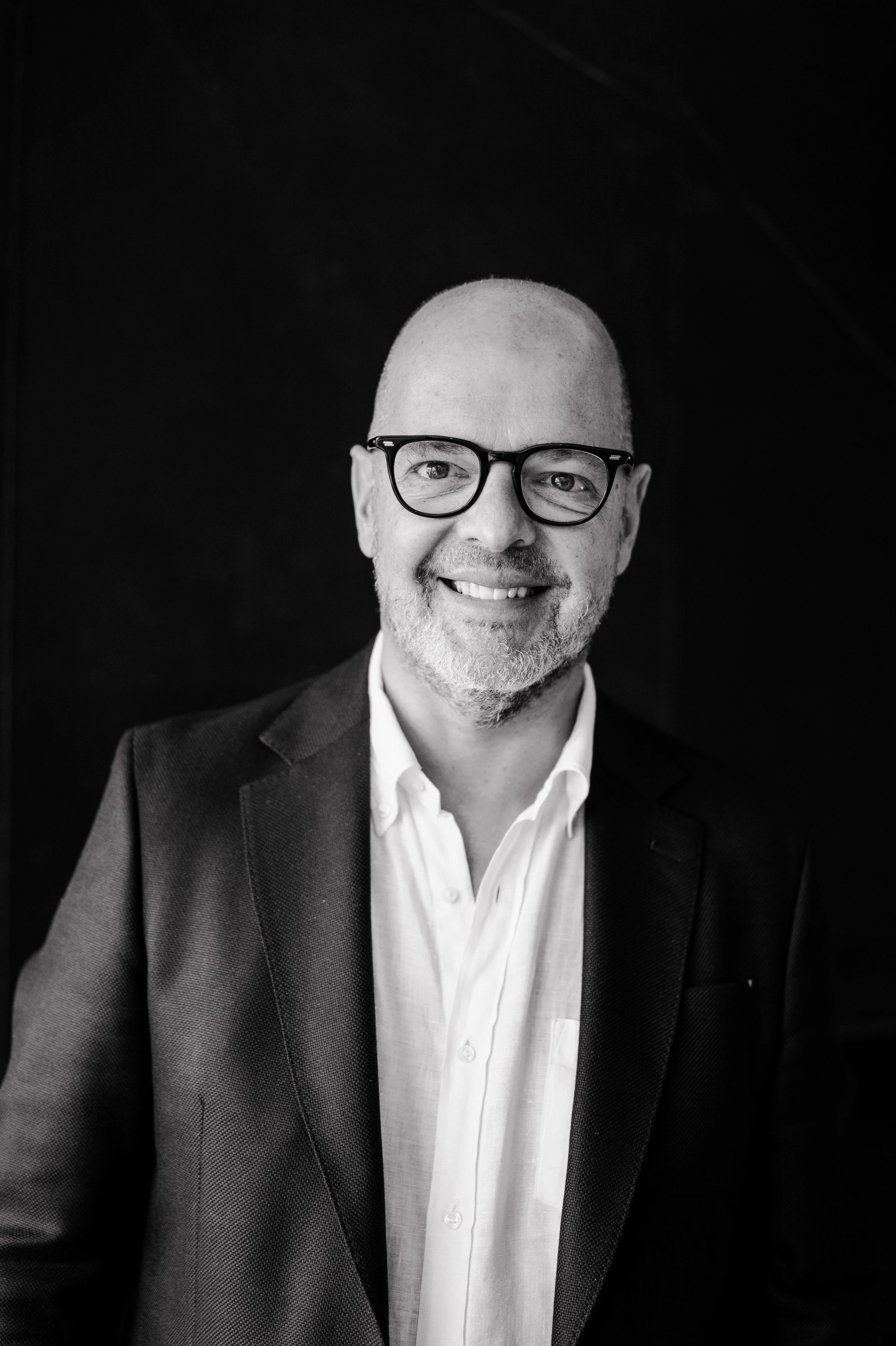 Black and white portrait of a smiling middle-aged man with glasses, a bald head, and a beard, wearing a suit and white shirt.