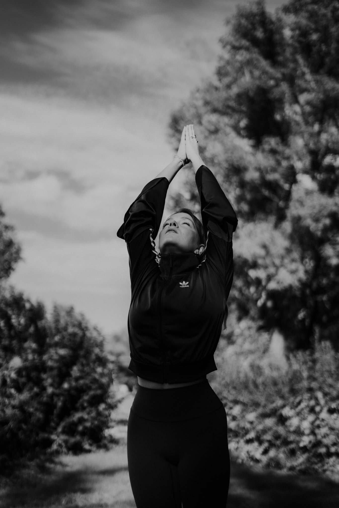 Schwarz-weiße Aufnahme einer Frau, die mit hochgereckten Armen in einer Yoga- oder Meditationshaltung steht, im Freien mit Bäumen im Hintergrund.