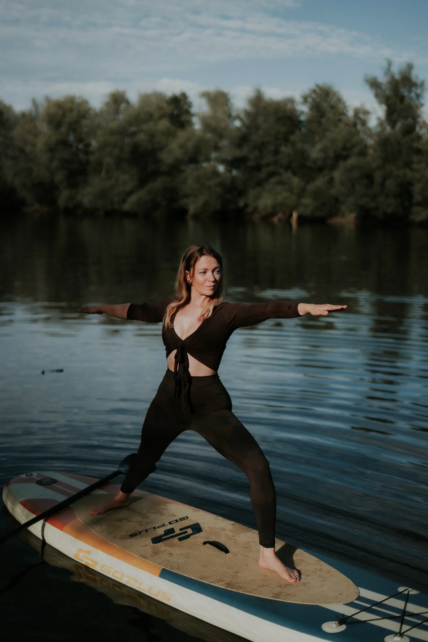 Eine Frau beim Yoga auf einem Stand-Up-Paddle-Board auf einem ruhigen See, mit Bäumen im Hintergrund und blauer Himmel.